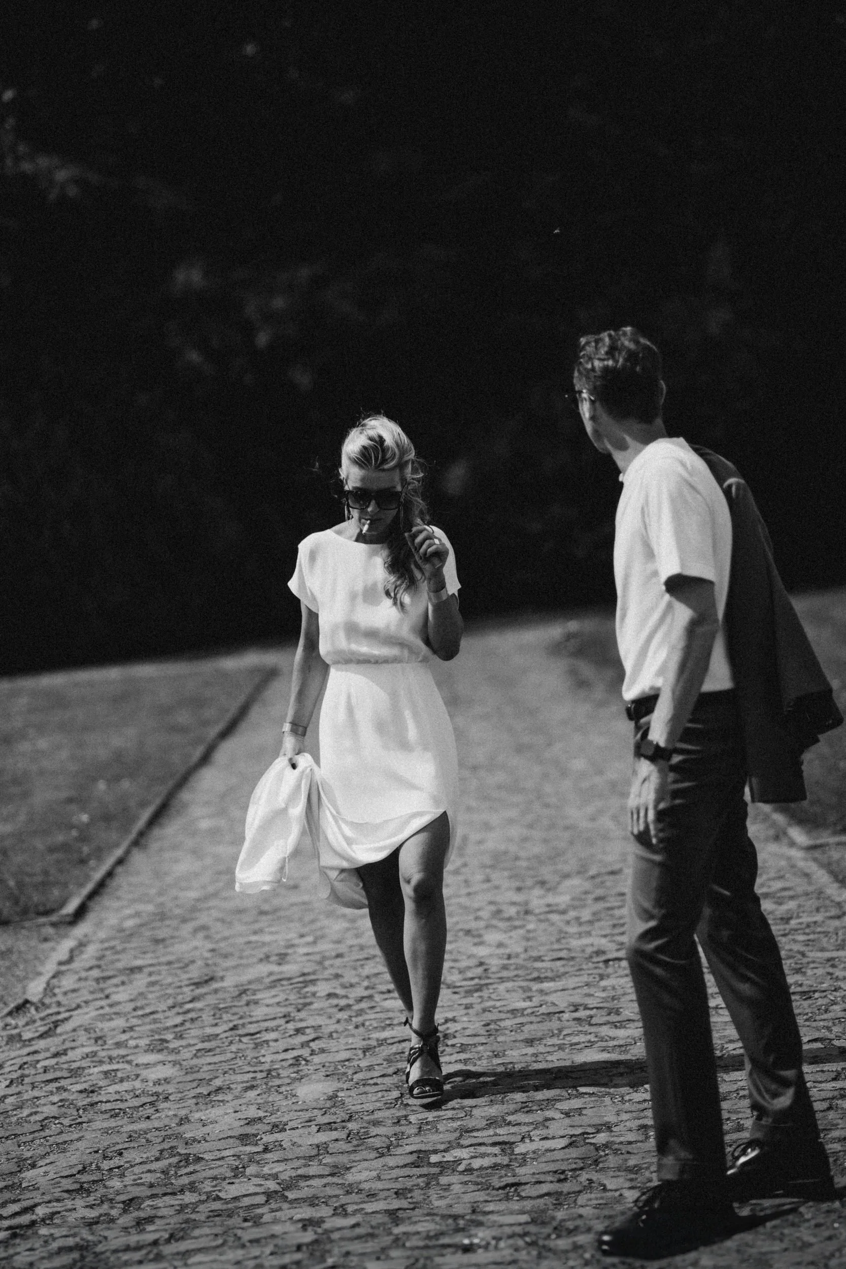A black and white photo of a woman in a white dress walking on a cobblestone path at night, with a man standing nearby, looking at her. Rockabilly style