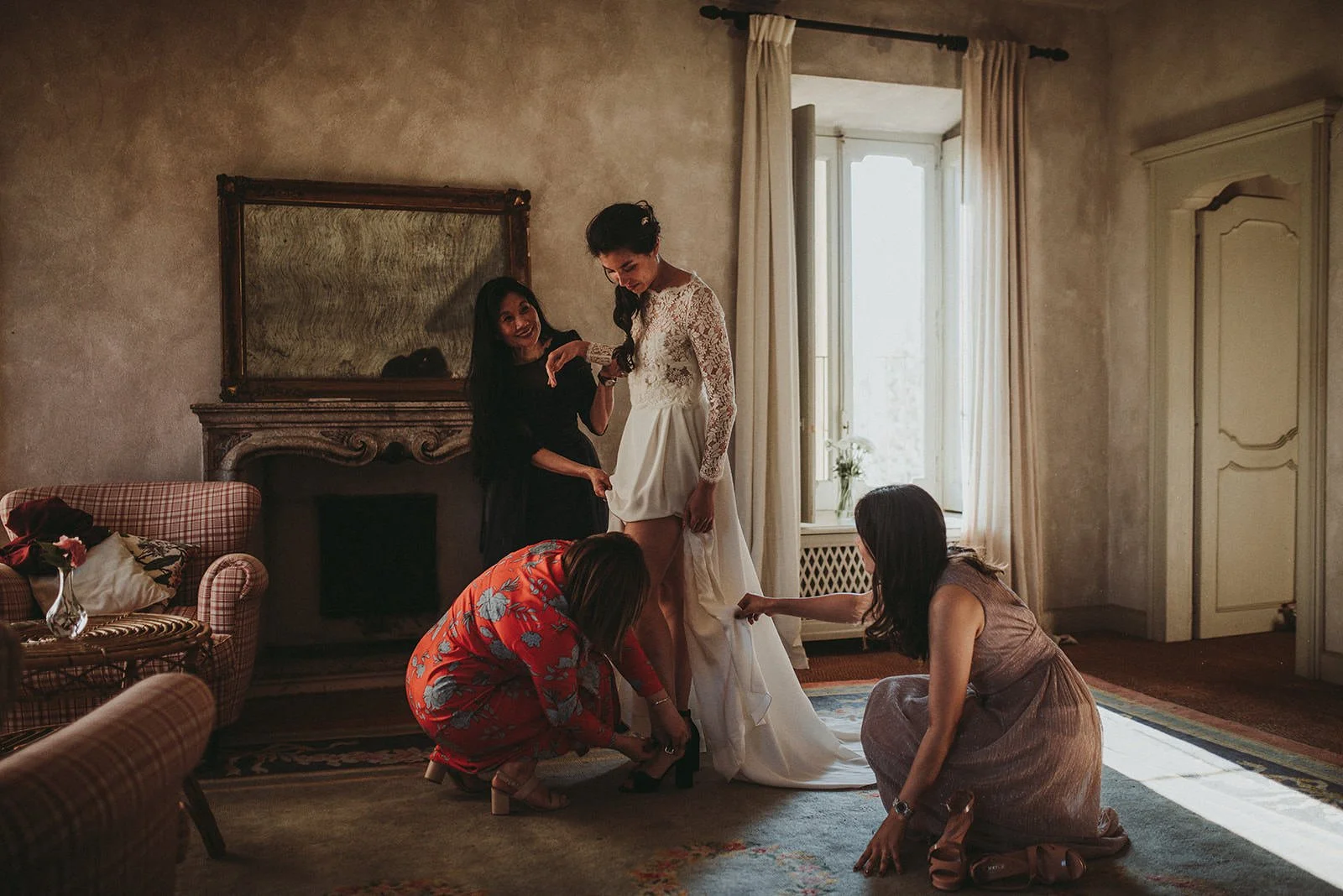 A bride in a wedding dress is standing with four women helping her with her shoes in a warmly lit room with a large mirror and window.
