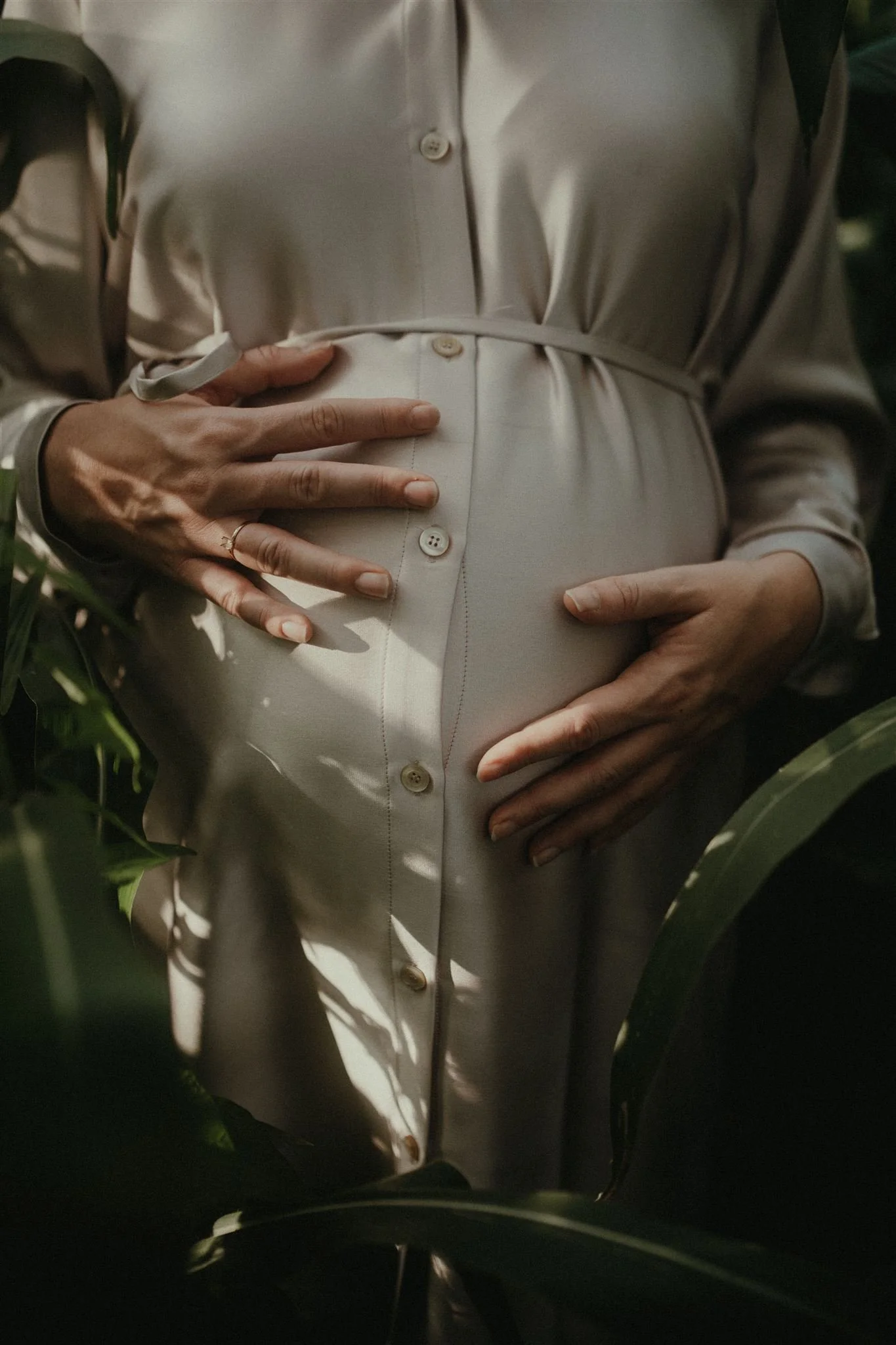 Close-up of a pregnant woman's belly being gently held by her hands, dressed in a beige button-up shirt, in a natural outdoor setting surrounded by green plants.