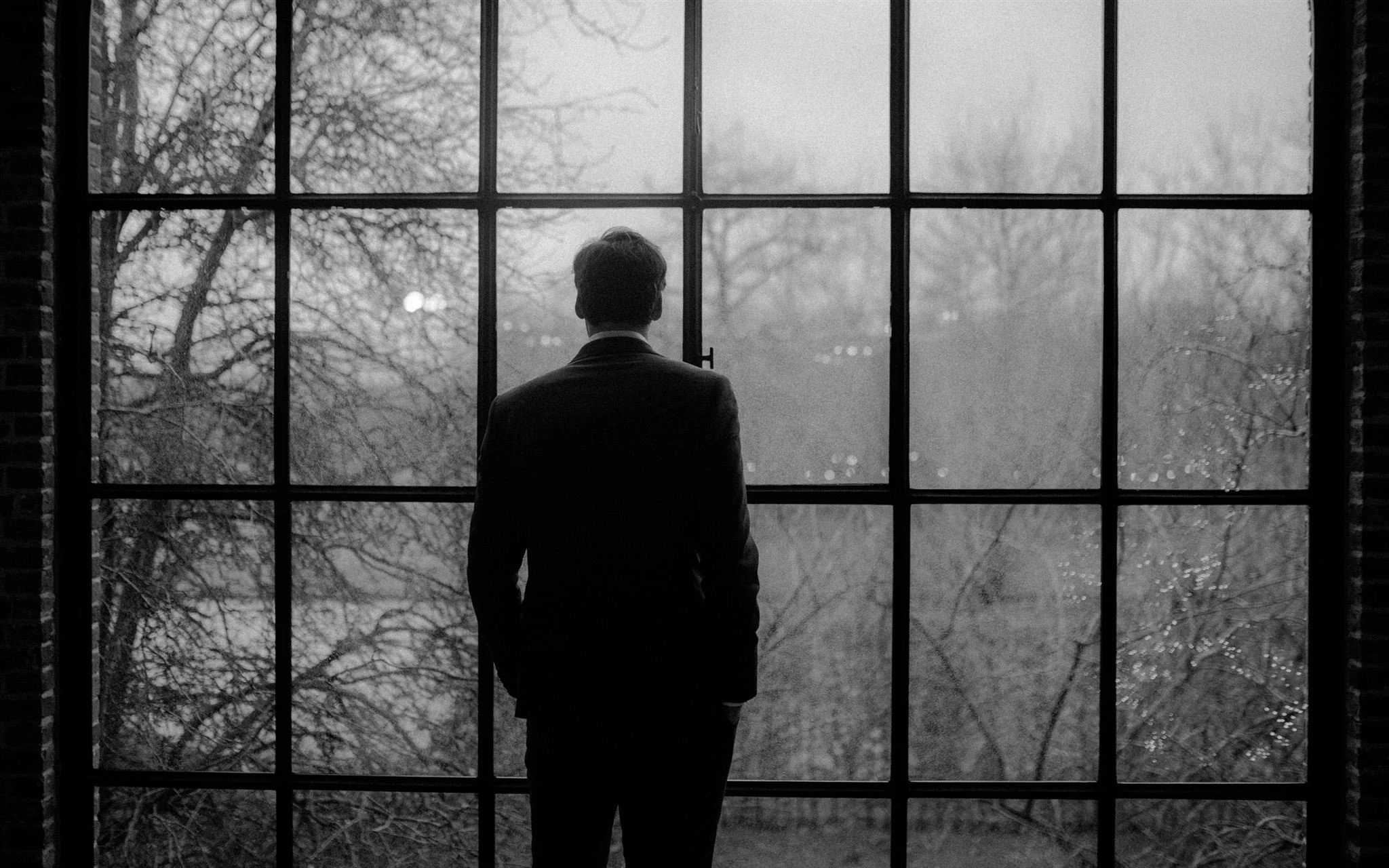 A man in a suit looking out a large window with a grid pattern at a winter scene with bare trees and a cloudy sky in black and white.