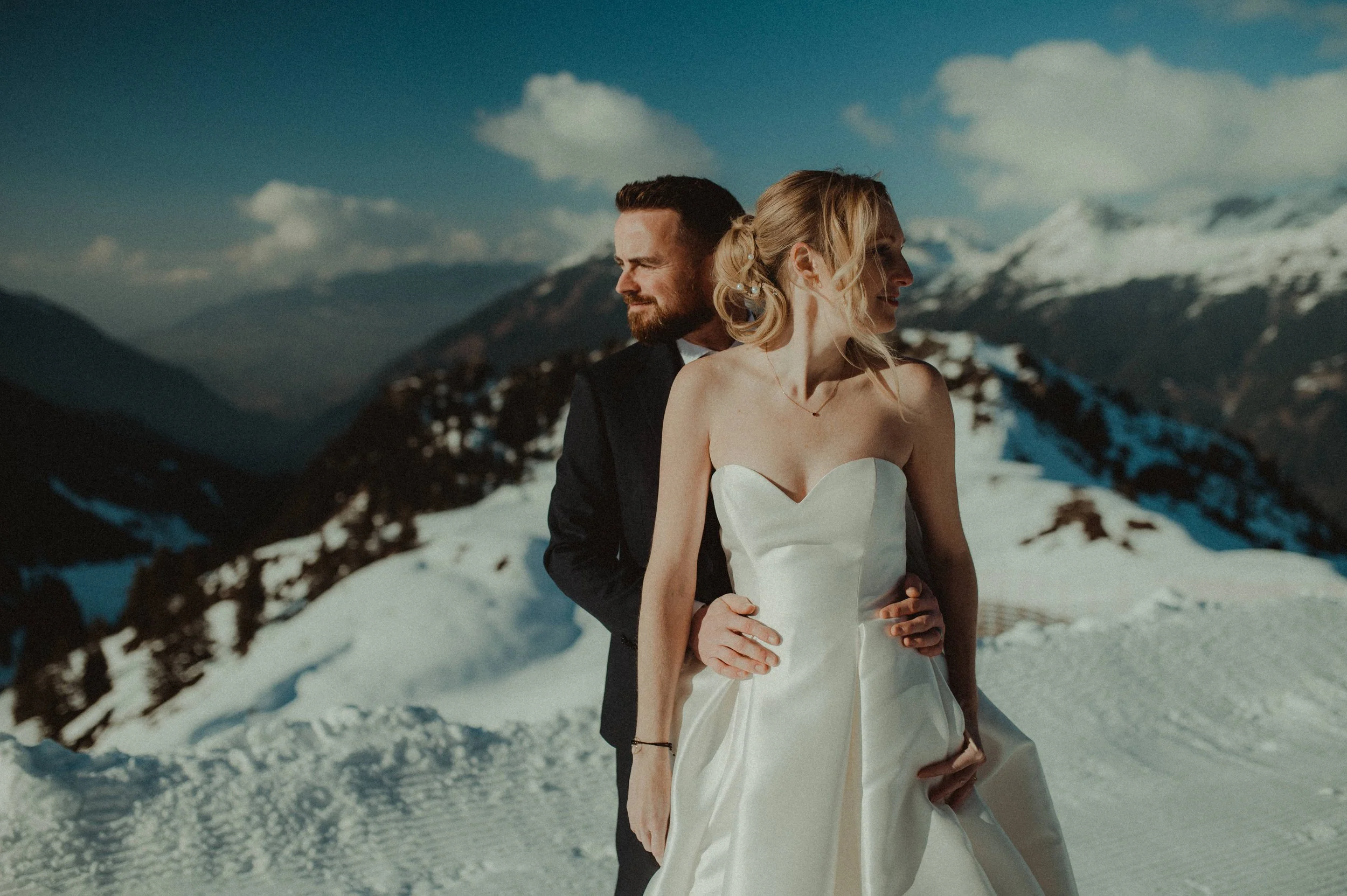 A couple in wedding attire standing on snow-covered mountains under a blue sky with clouds, embracing each other.