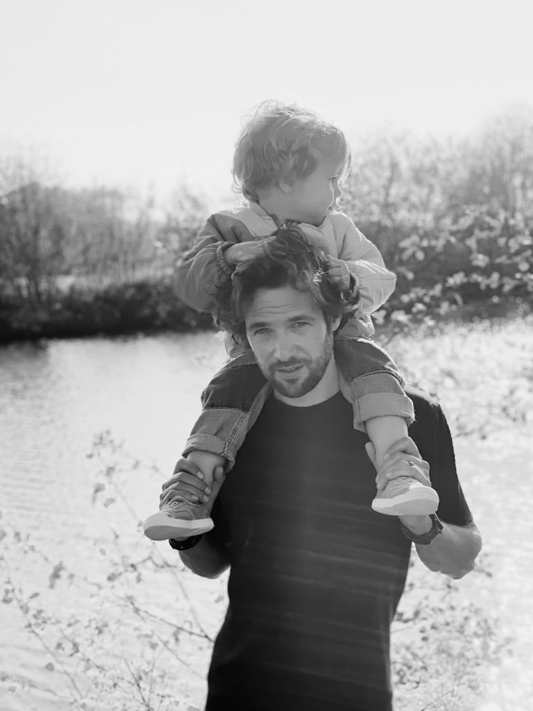 A man carrying a young child on his shoulders outdoors near a body of water with trees in the background.