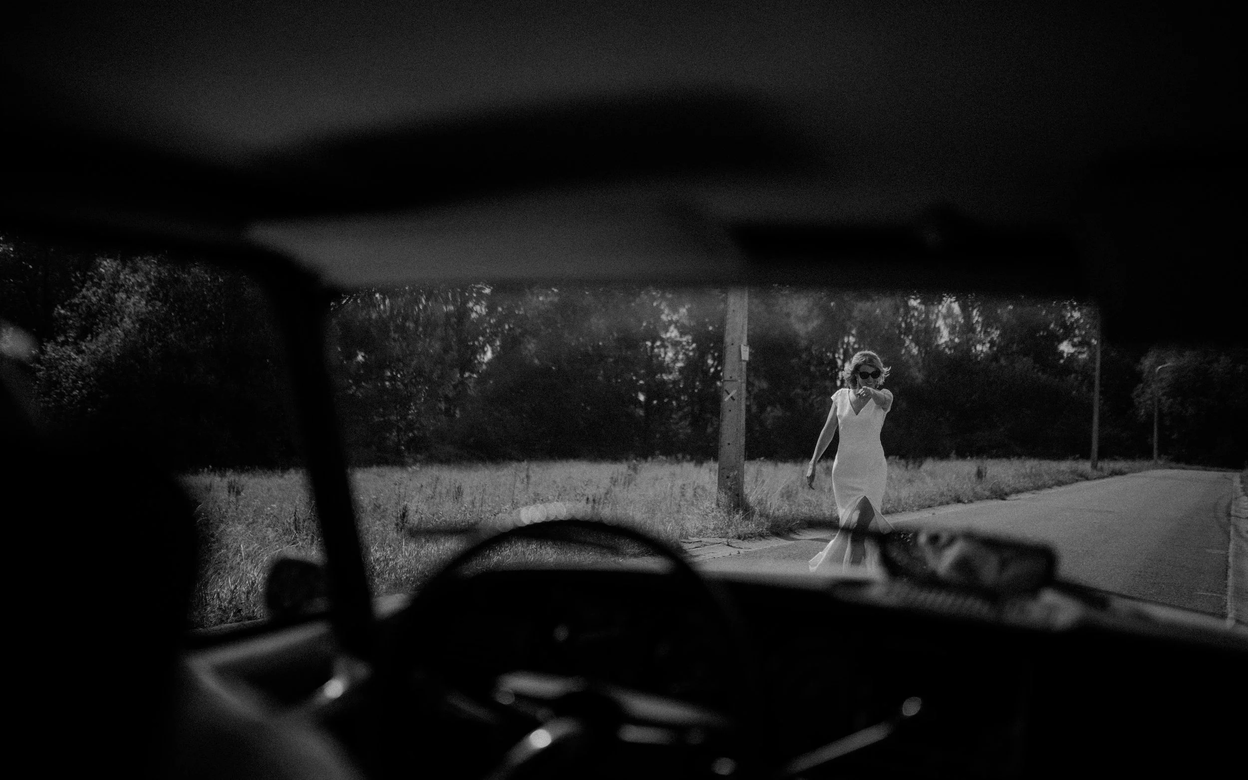 Black and white photo of a woman walking on a rural road viewed from inside a car. The woman is wearing a white dress, sunglasses, and has curly hair, walking towards the car.