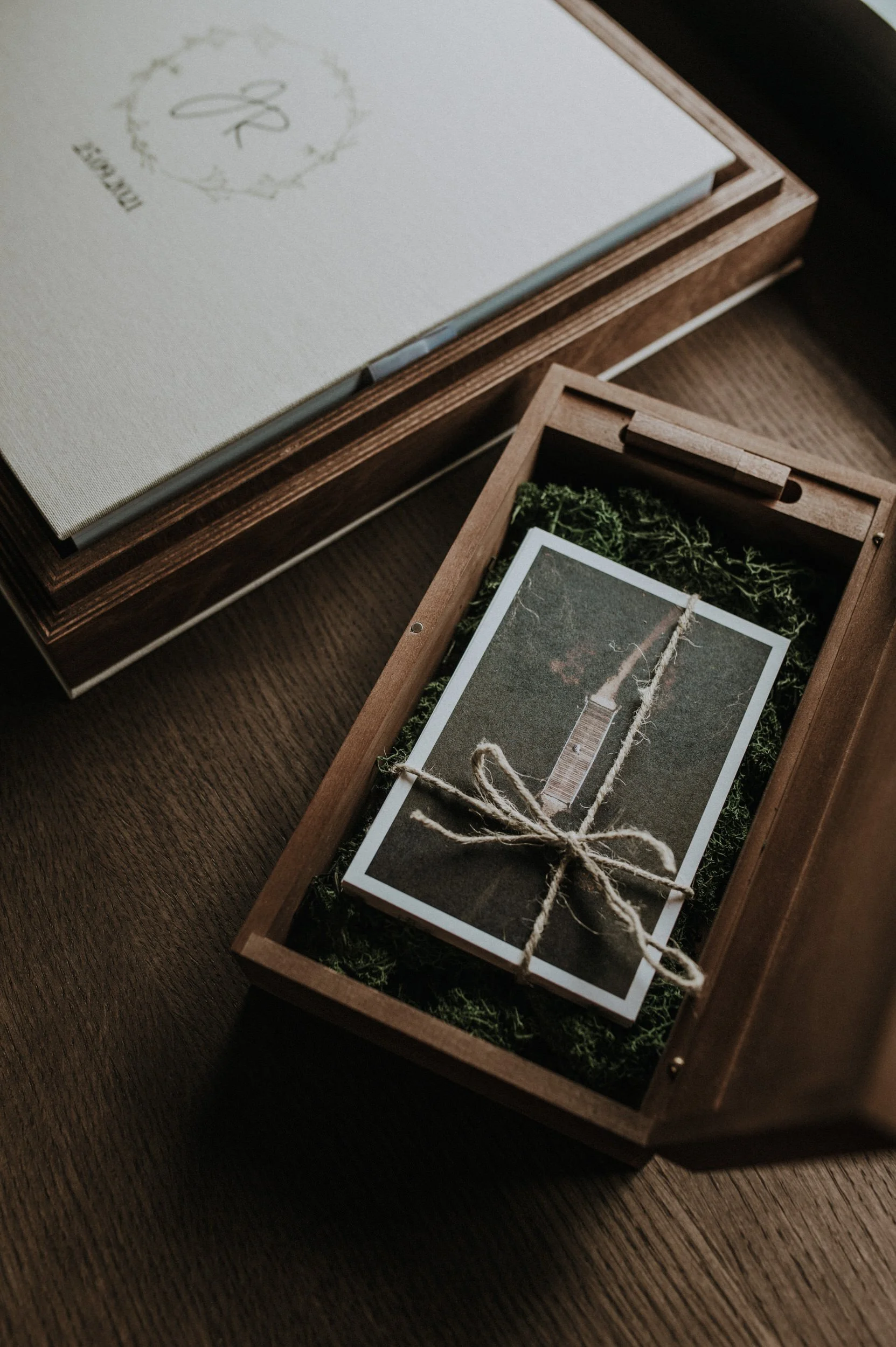 A wooden box with a photo of a dock on it, tied with twine, inside a bed of green moss, alongside a closed white box with a monogram and decorative border.