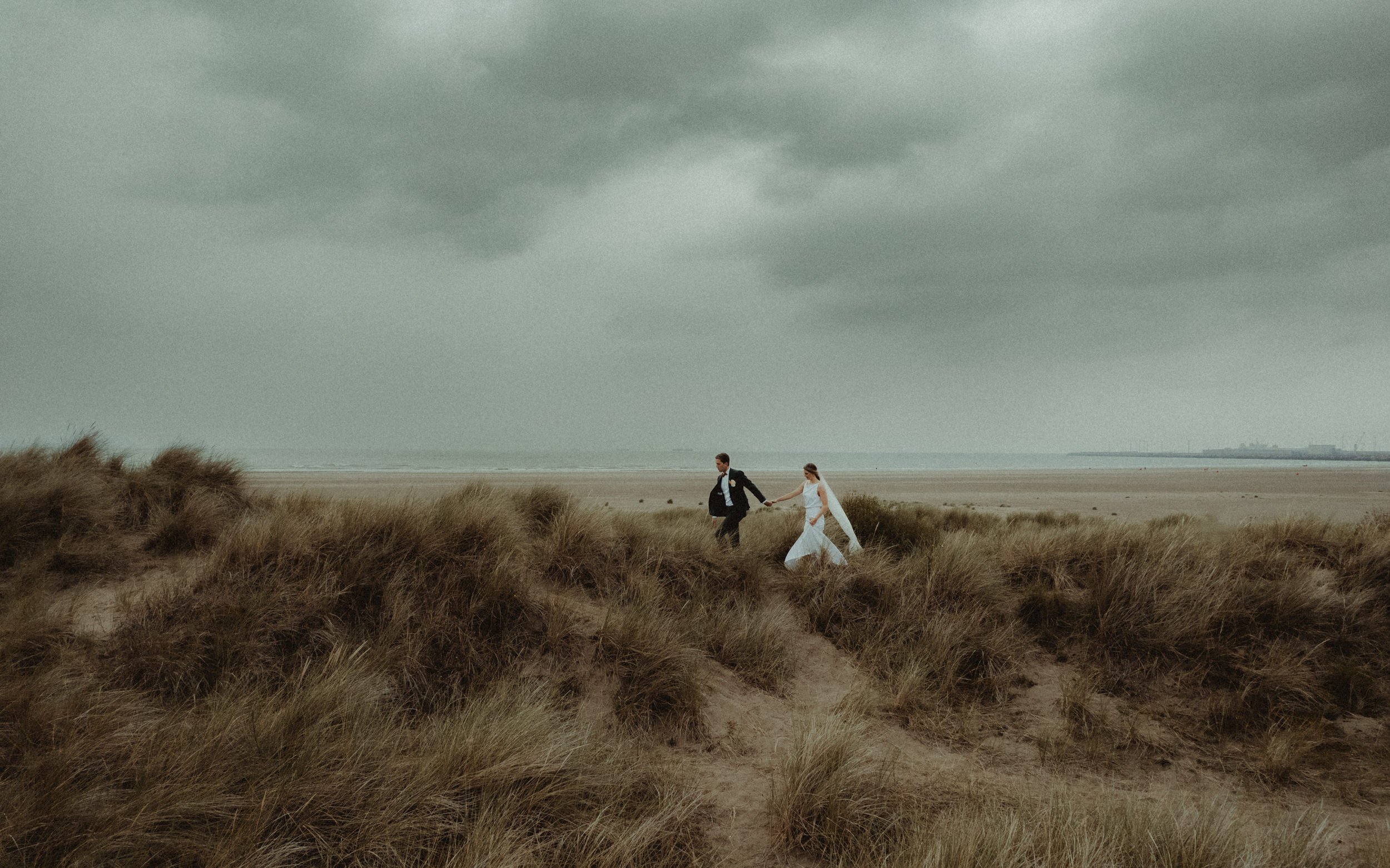 A couple dressed in wedding attire walking hand-in-hand through sand dunes on a cloudy beach.