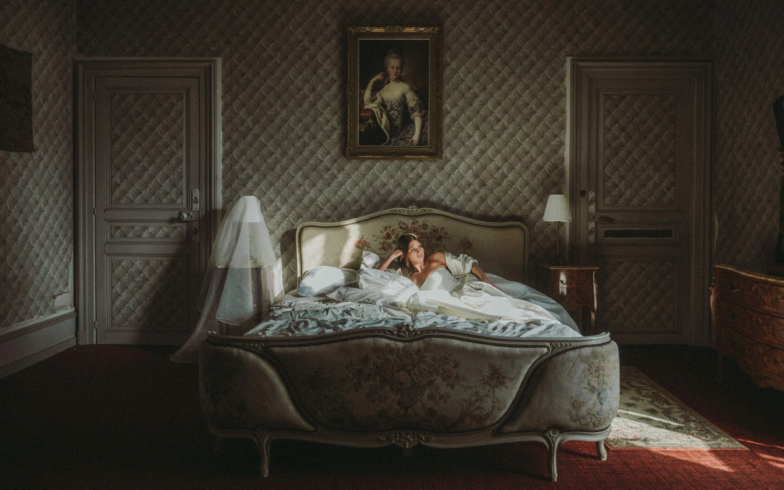 A woman lying on a vintage bed in a dimly lit bedroom with ornate furniture and patterned wallpaper, sunlight streaming through a window on the right.