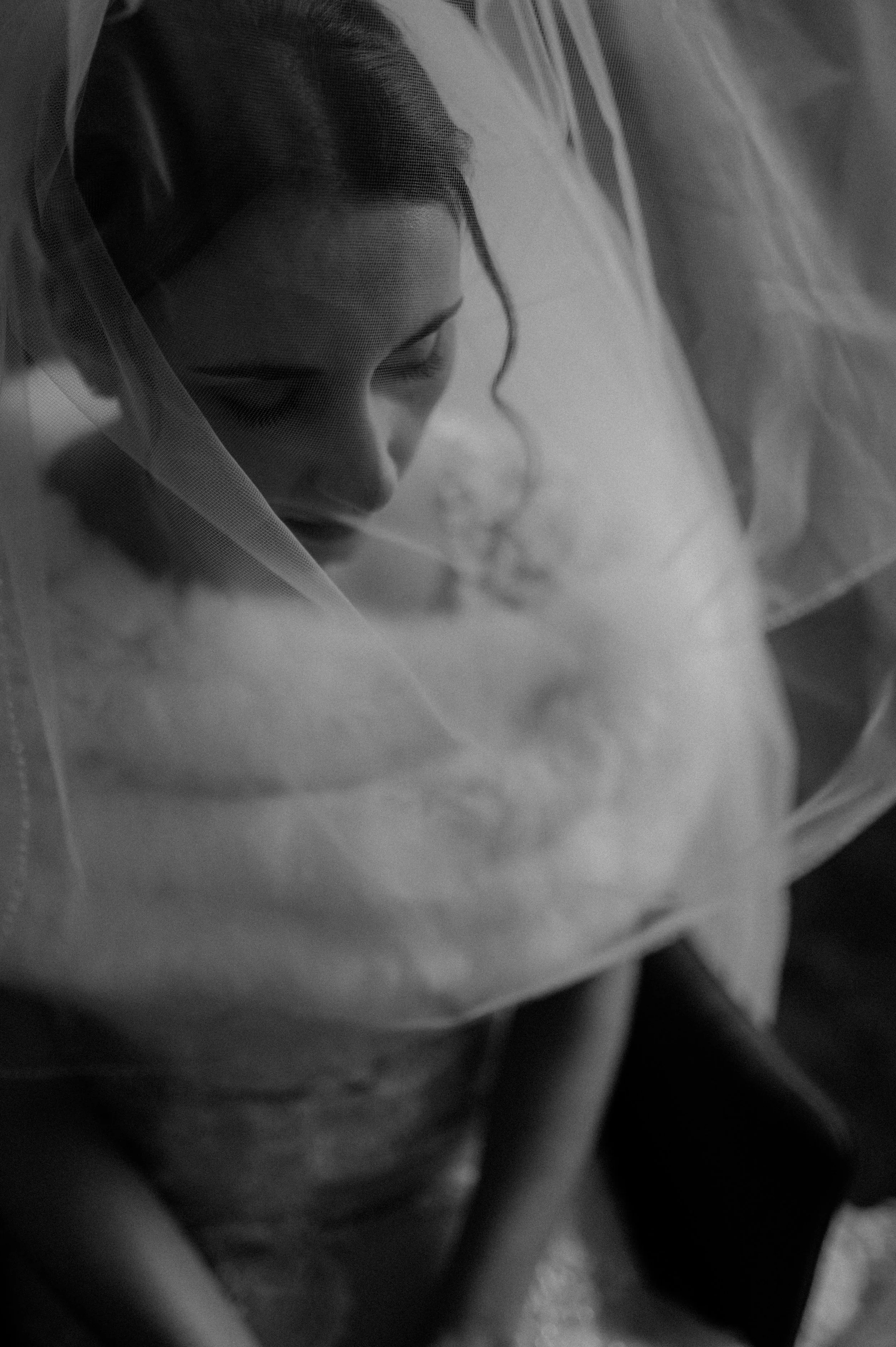A woman with dark hair and makeup, wearing a sheer veil over her face and shoulders, appears in a contemplative pose in a black and white photograph.
