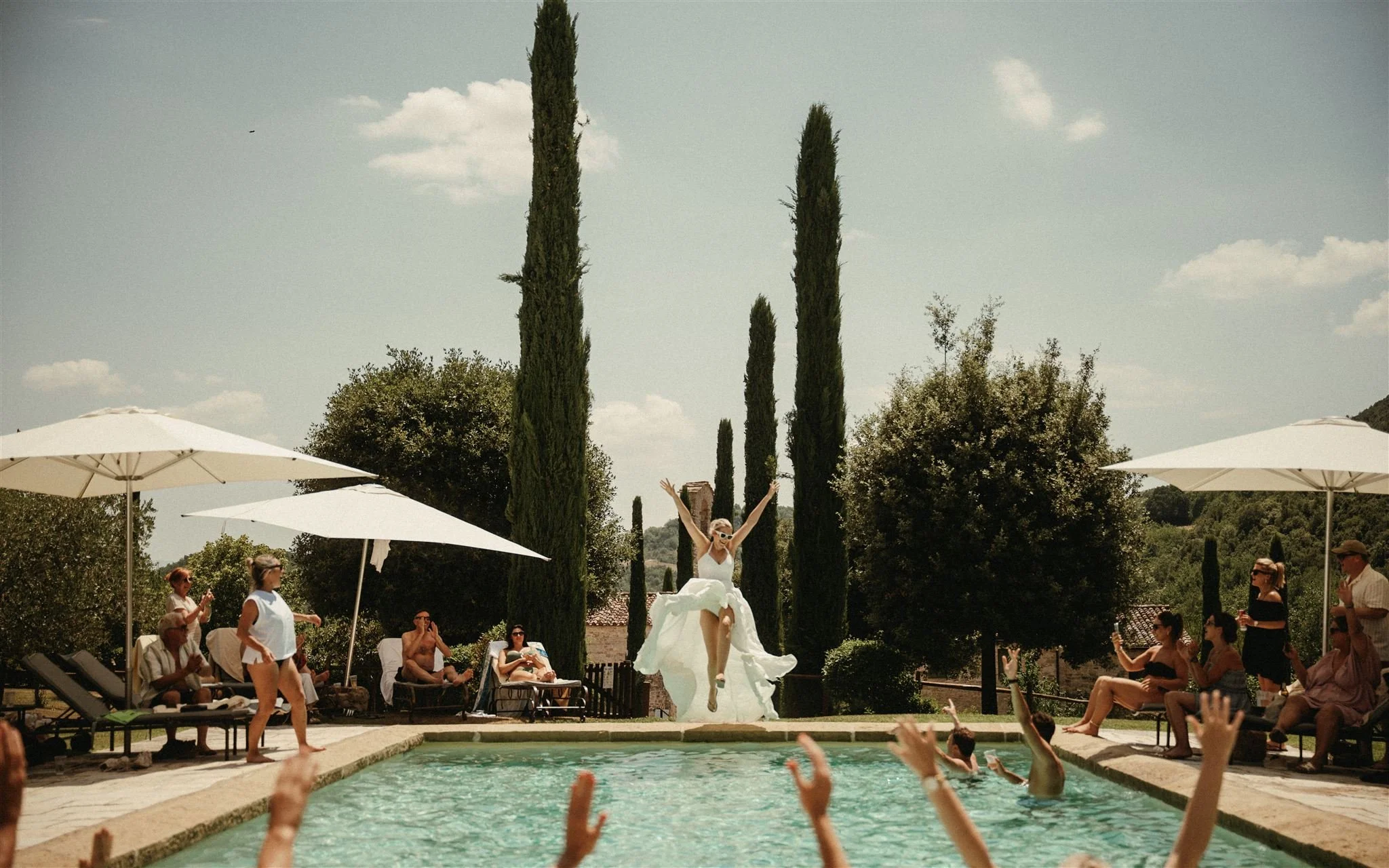A woman in a wedding dress jumps into a swimming pool during a celebration, while guests watch and take photos around the pool area on a sunny day.
