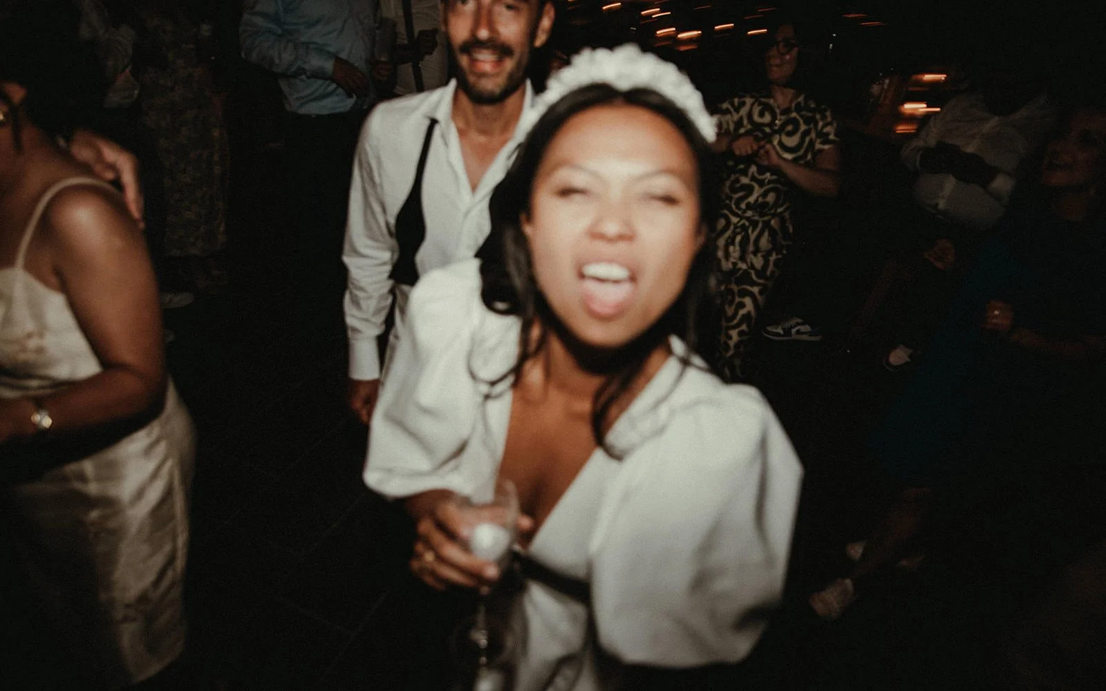 A woman in the foreground wearing a white headband and white dress makes a playful face at a lively social gathering or party, holding a drink in her hand. Others are seen in the background, smiling and mingling.