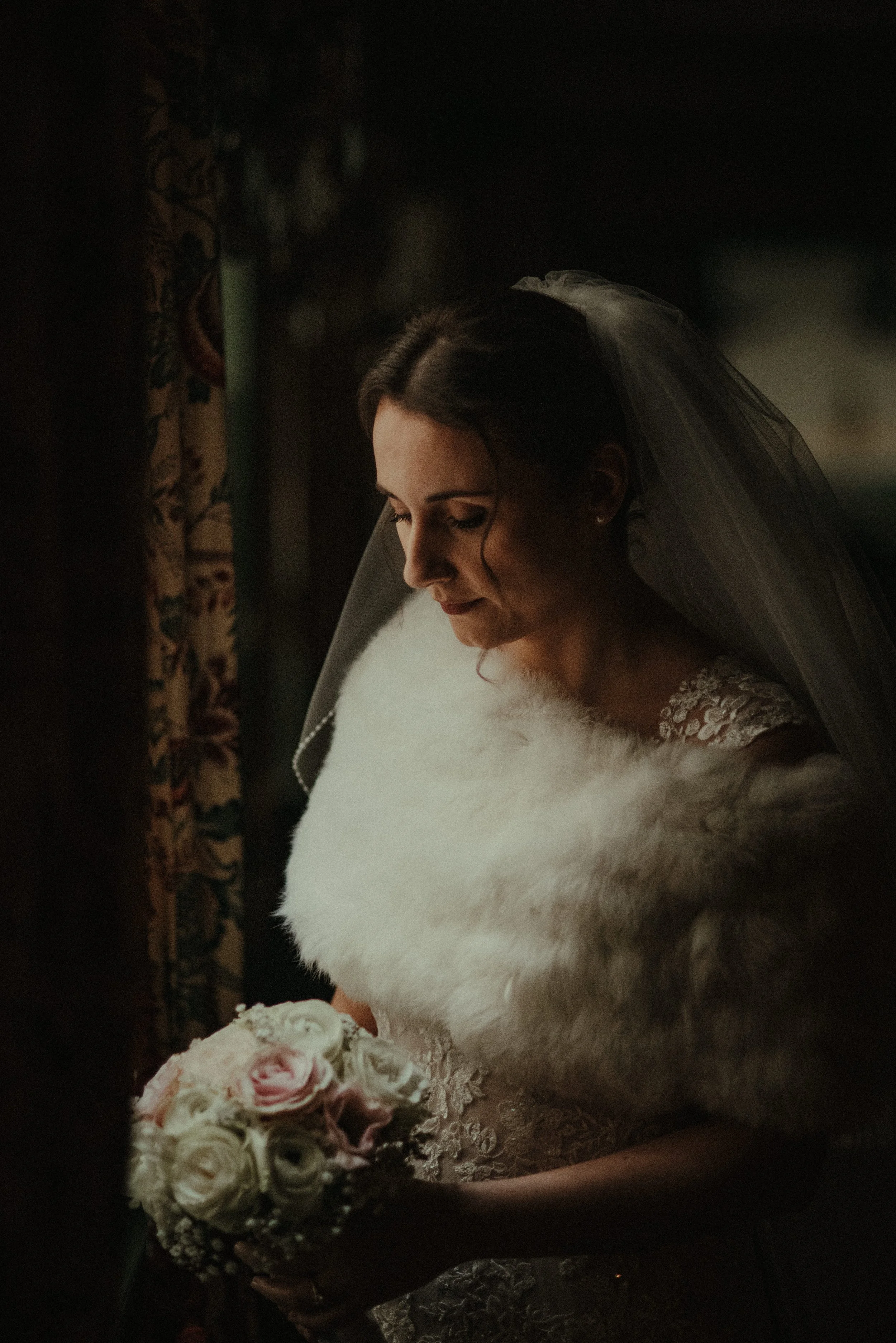 Bride in a wedding dress with a white lace bodice, wearing a veil and holding a bouquet of pink and white roses, standing near a window with dark curtains.