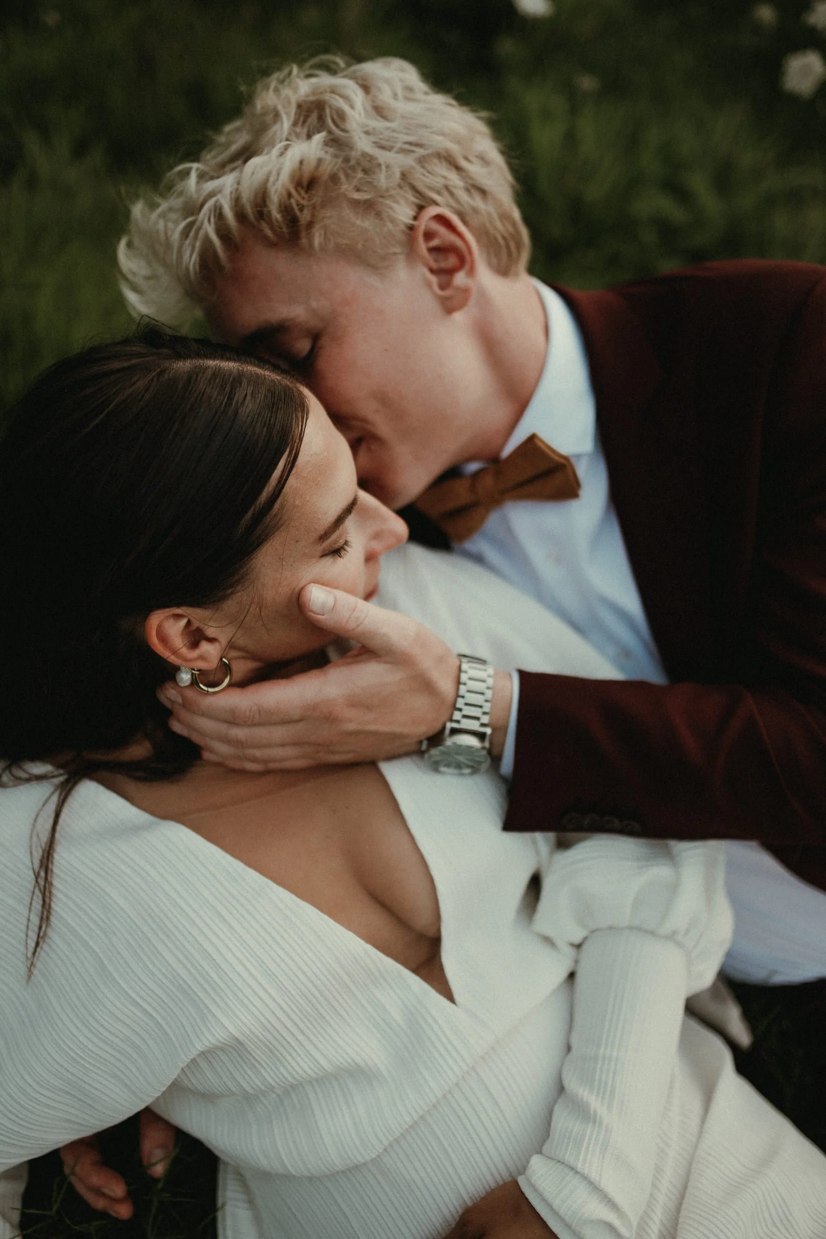 A couple lying on the grass, sharing an intimate moment. The man, with curly blond hair, is leaning over and gently holding the woman's face. The woman, with dark hair and pearl earrings, is smiling softly.