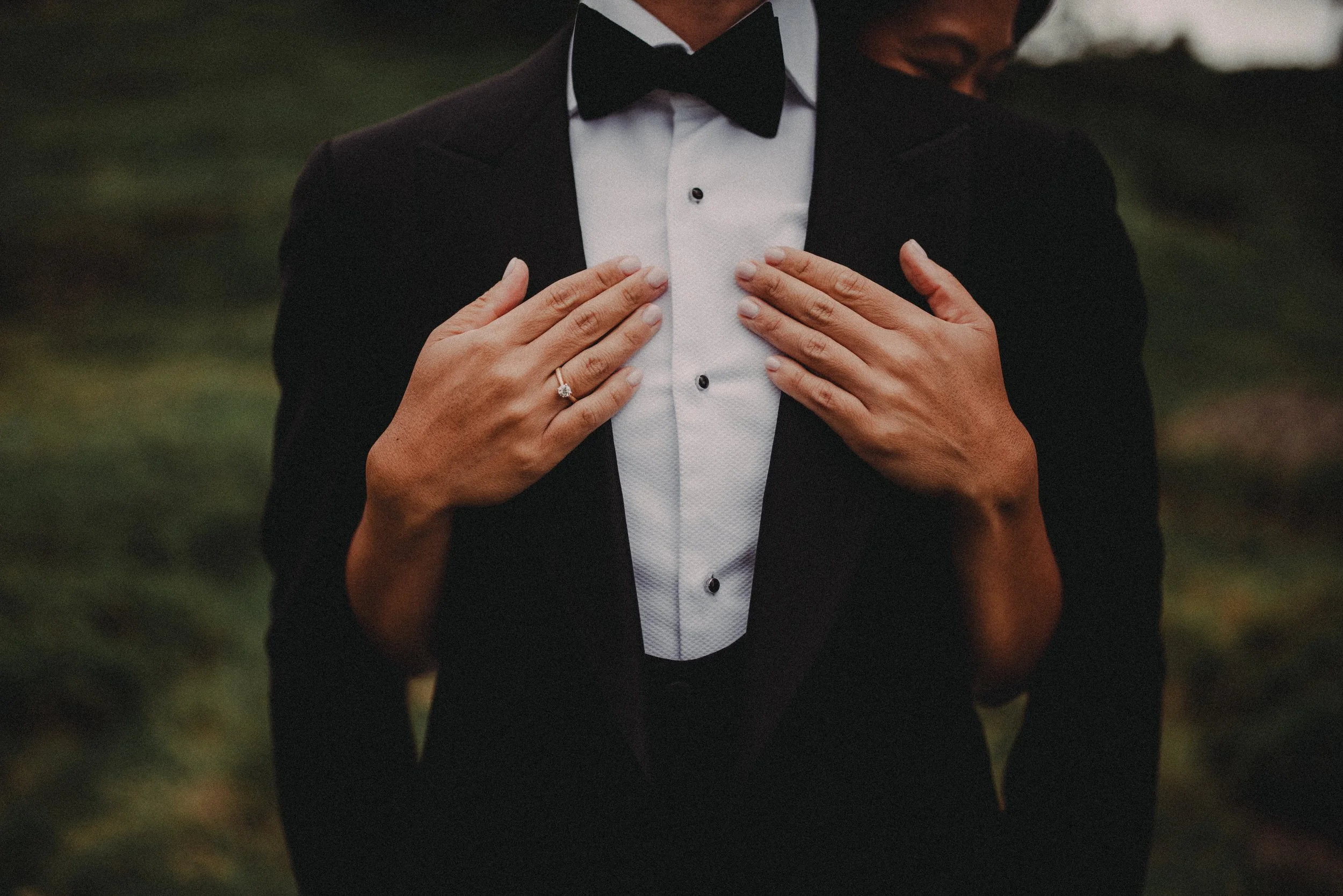 A person in a tuxedo with a white shirt and black bow tie is holding their hands over their chest, showing a wedding ring on their left hand.