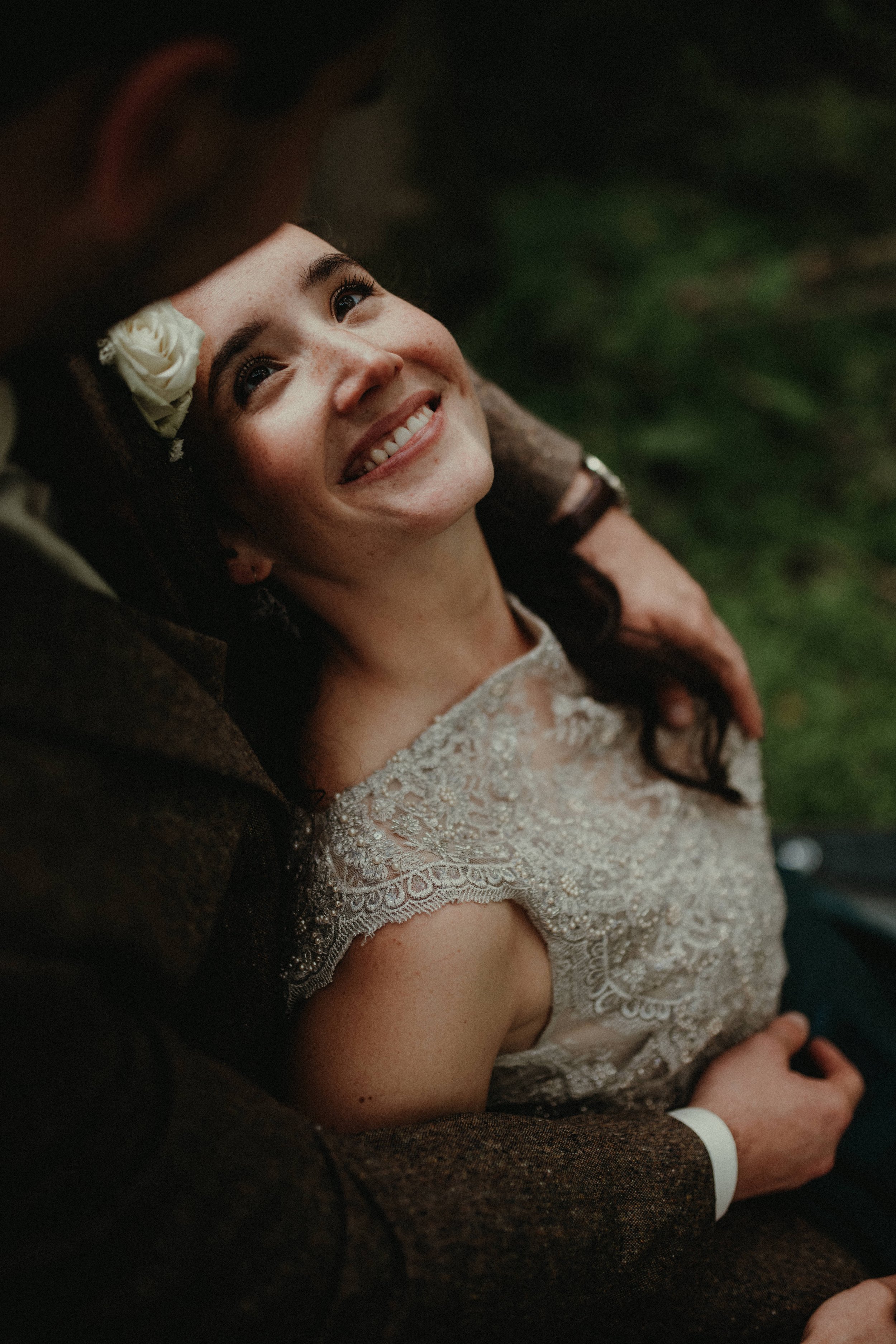 Close-up of a smiling woman with dark hair and freckles, wearing a lace dress, lying on a person's lap, with a flower in her hair and looking up at them.