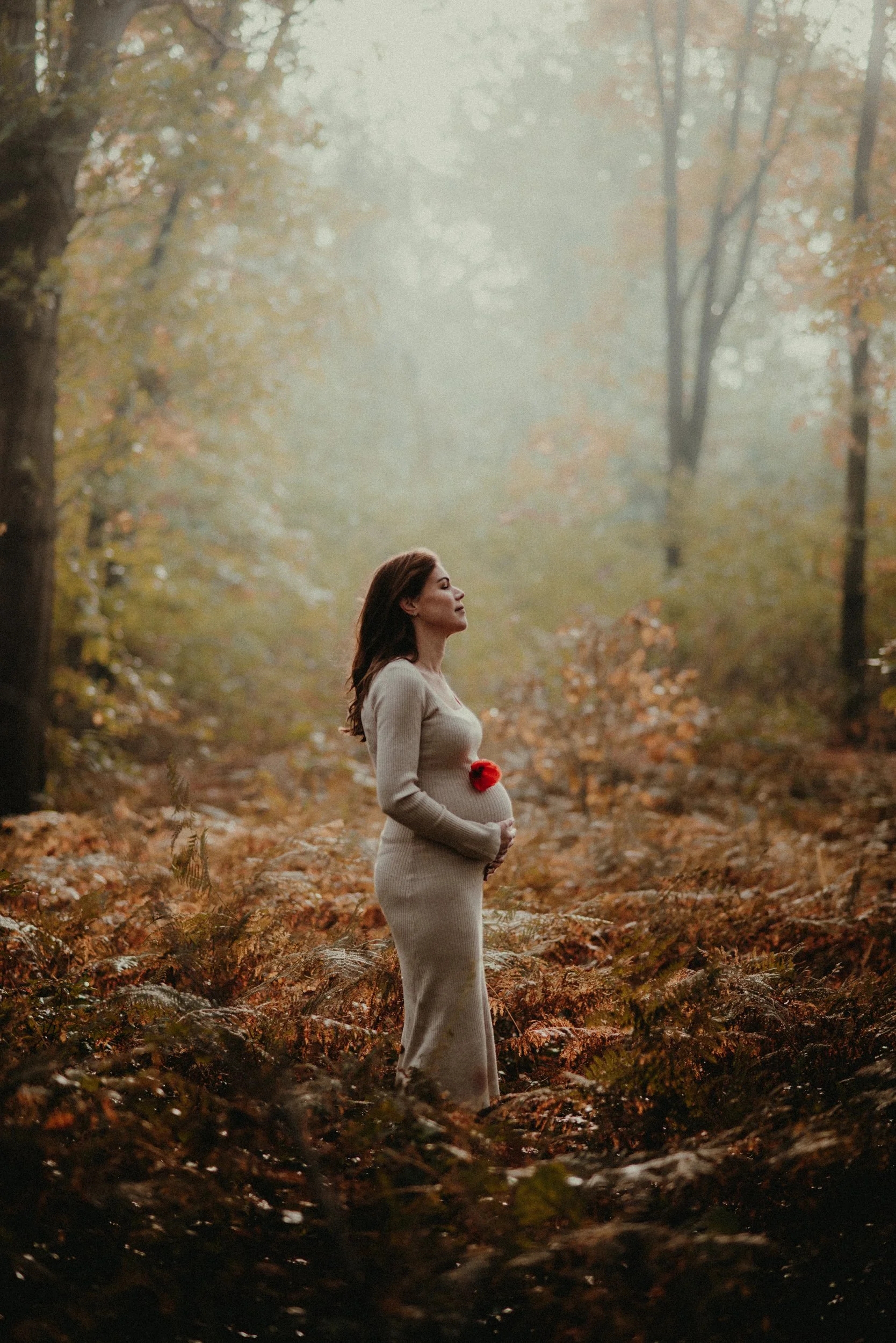A pregnant woman in a beige dress standing in a foggy forest during autumn, holding her belly with a red flower pinned to her dress.