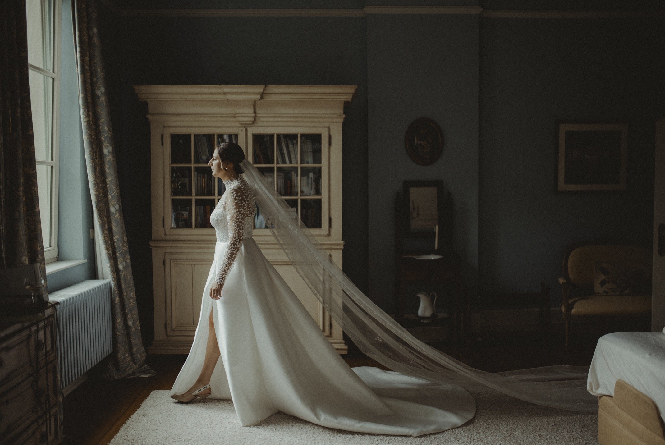 A bride in a wedding dress with lace sleeves stands indoors near a window, illuminated by natural light.