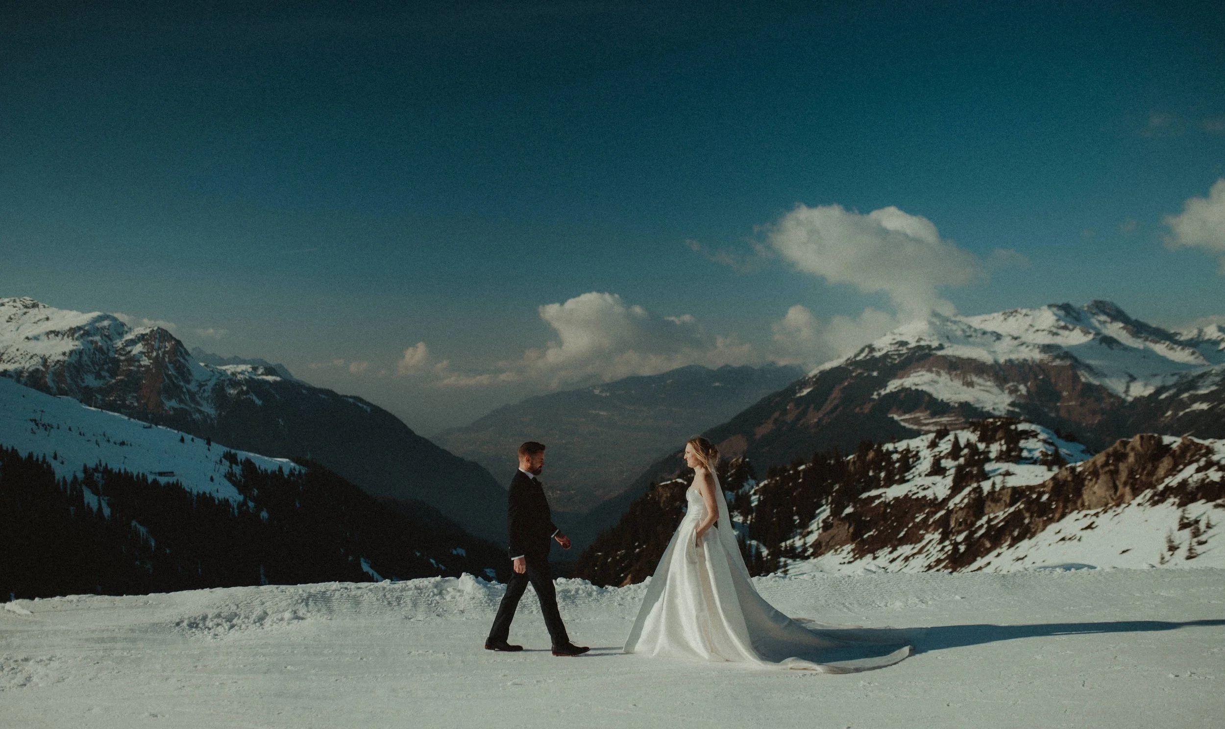 A bride and groom standing on snow-covered ground in a mountainous landscape with snow-capped peaks under a partly cloudy sky.