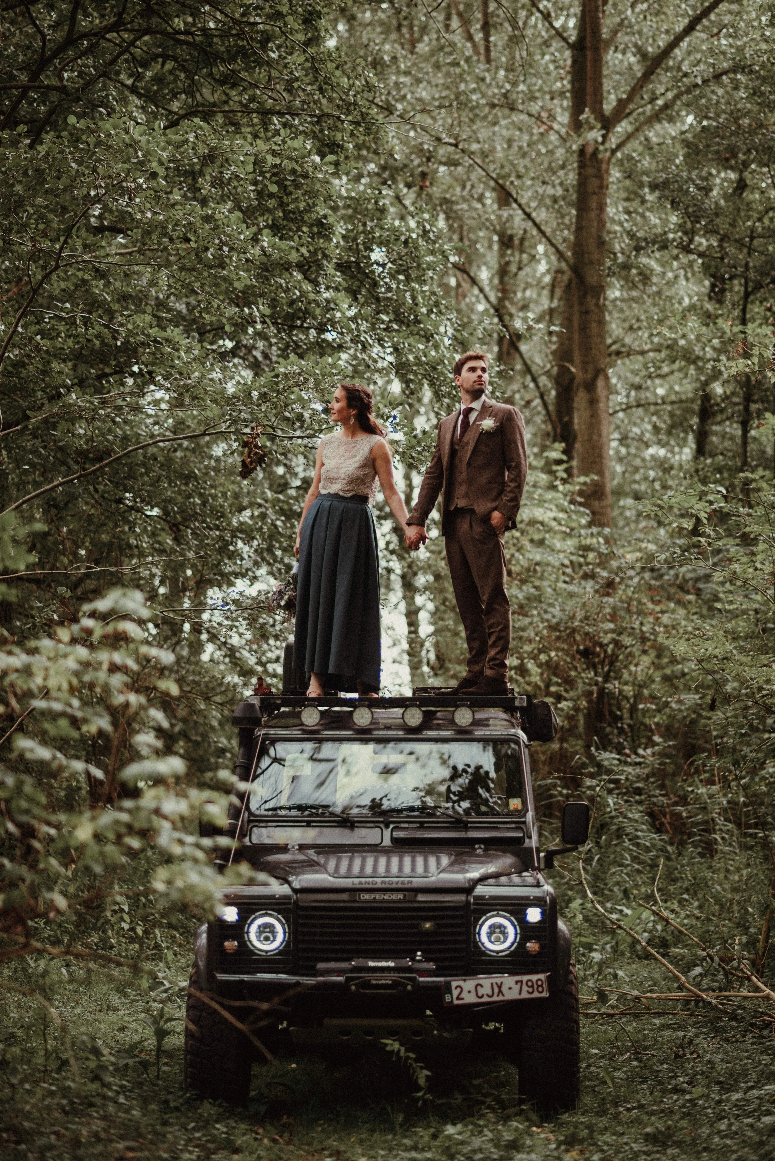 A couple dressed in vintage wedding attire standing on top of a black Land Rover in a dense forest, holding hands and gazing into the distance.