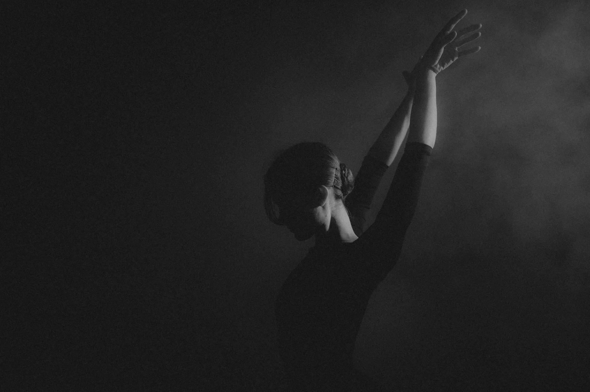 A woman with short hair and a dark top is raising her arms gracefully in a dance pose, with dramatic lighting creating shadows and highlights on her face and body against a dark background.