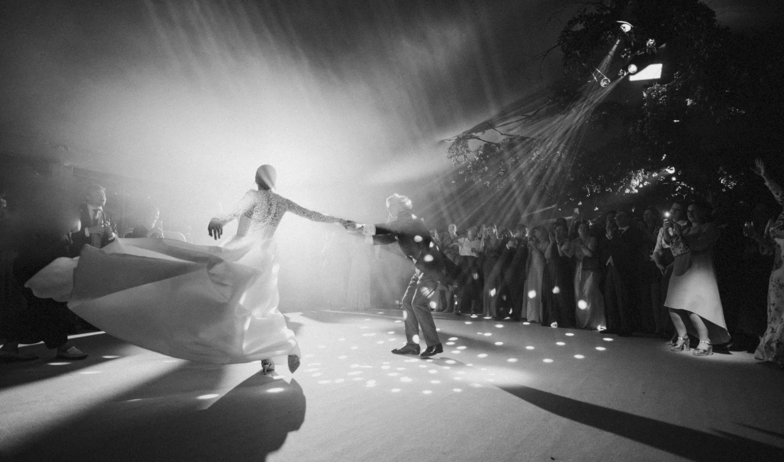 A black-and-white photo of a lively dance scene at a celebration, with a woman in a flowing dress spinning on the dance floor, surrounded by onlookers capturing the moment with their phones, and dramatic lighting creating a festive atmosphere.