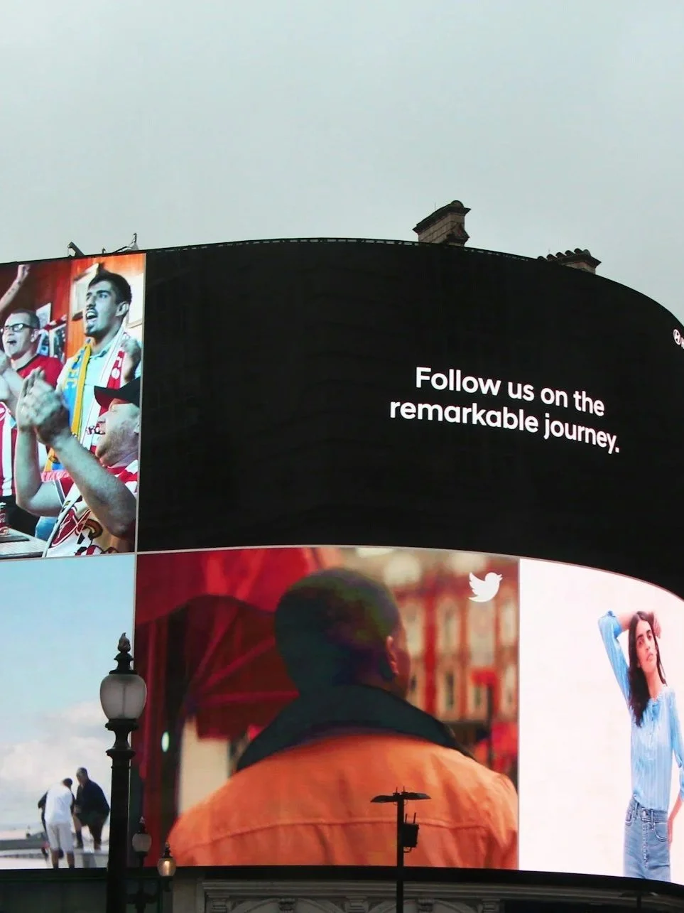 A large digital billboard showing the message "Follow us on the remarkable journey" along with various images of diverse people, including a group of sports fans and individuals in outdoor settings.