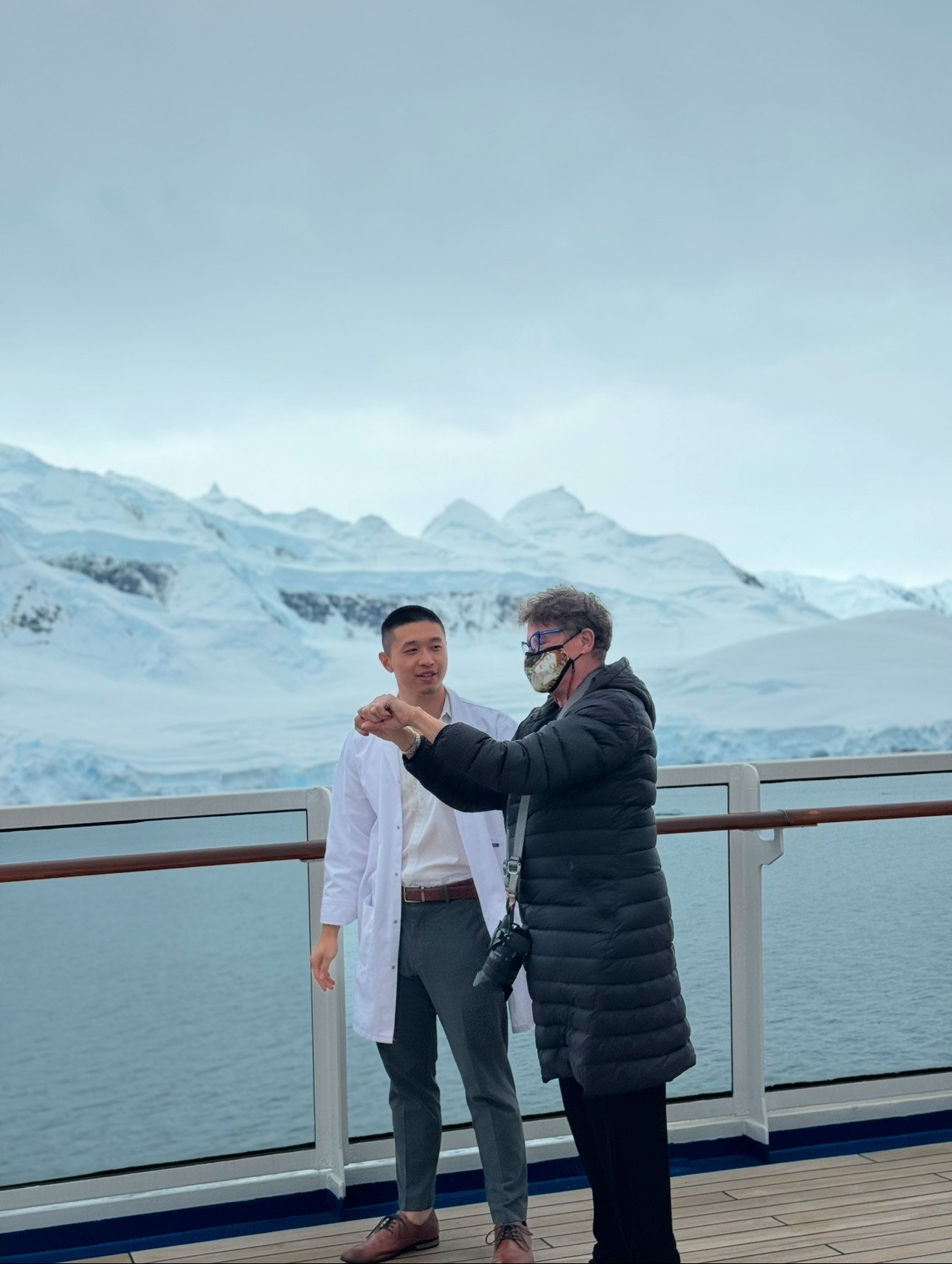 A patient joyfully showing improved hand mobility after acupuncture, standing with The Gentle Needle Guy on a ship deck in Antarctica with snowy mountains in the background.