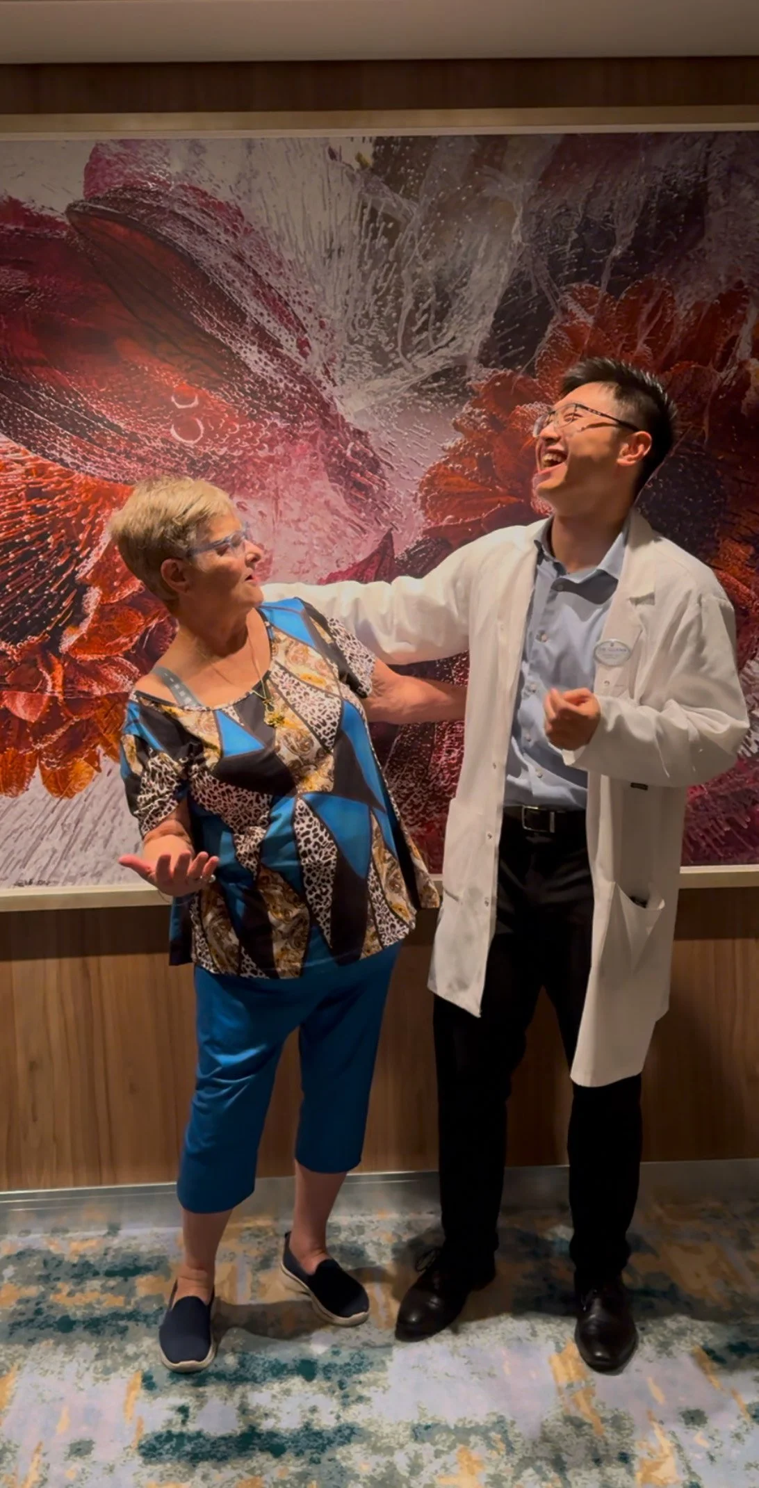 Two people laughing in front of a colorful abstract painting — The Gentle Needle Guy in a lab coat and a patient expressing joy and relief after experiencing pain reduction through acupuncture.