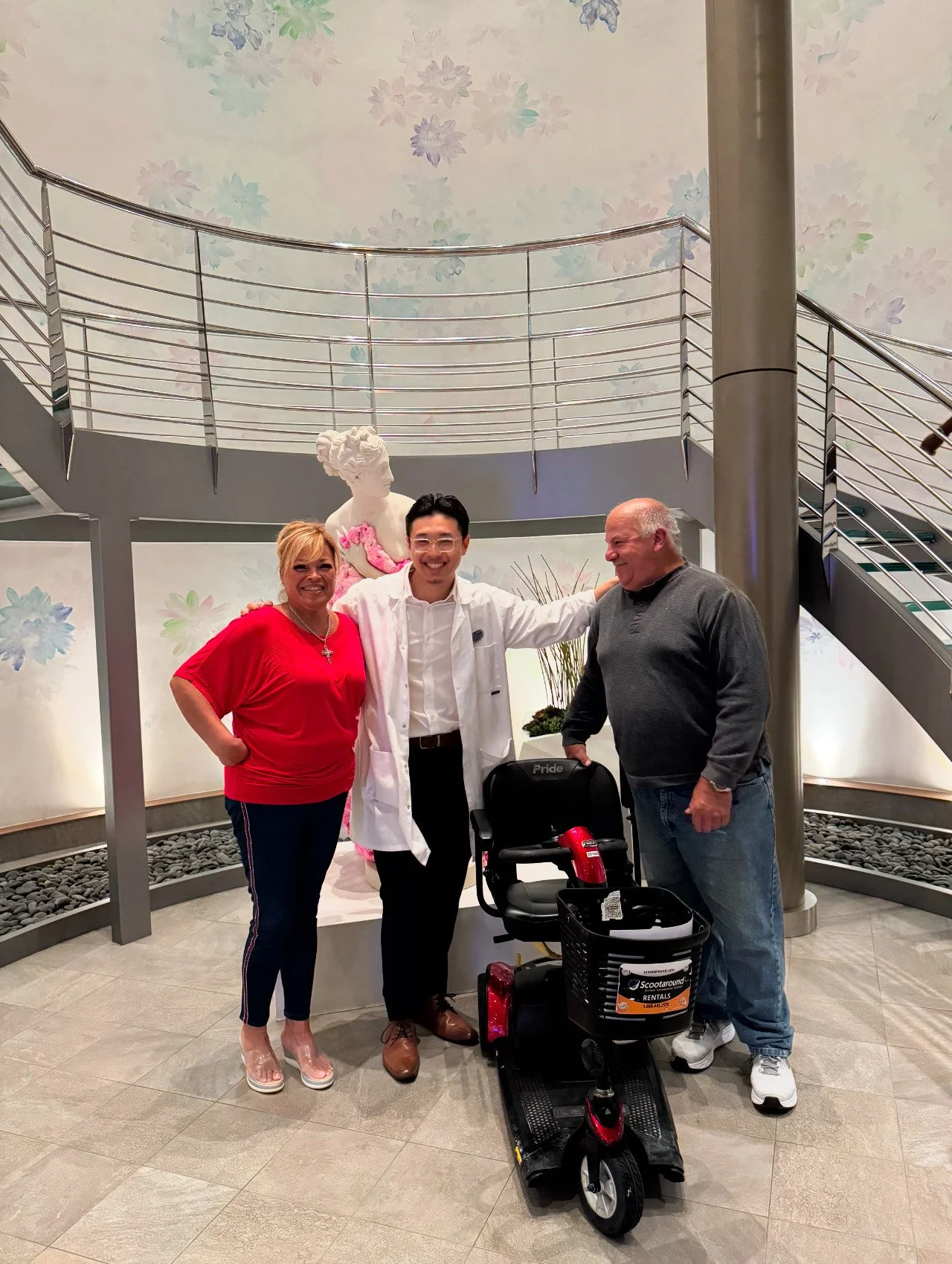 Three people standing inside a building with a spiral staircase in the background. A man in a white coat is in the middle, a woman in a red outfit on the left, and an older man in casual clothing on the right next to a mobility scooter.