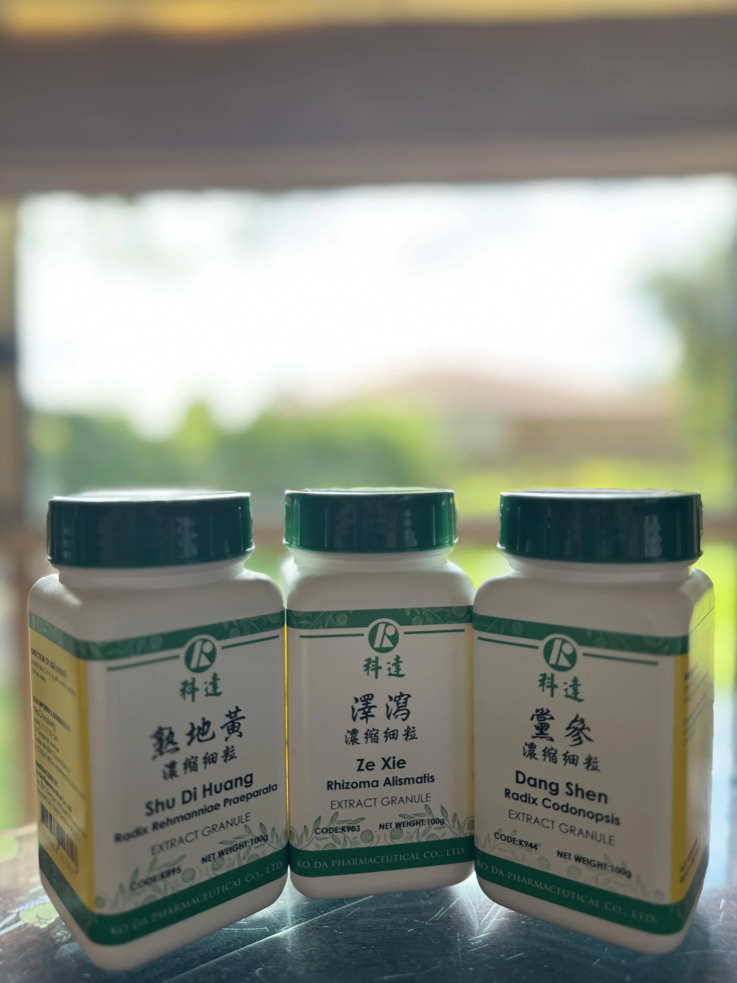 Three bottles of herbal extract supplements with green caps, labeled in Chinese and English, placed on a blue surface with a blurred window view — reflecting the integrative herbal approach at The Gentle Needle Guy’s clinic in Brisbane.