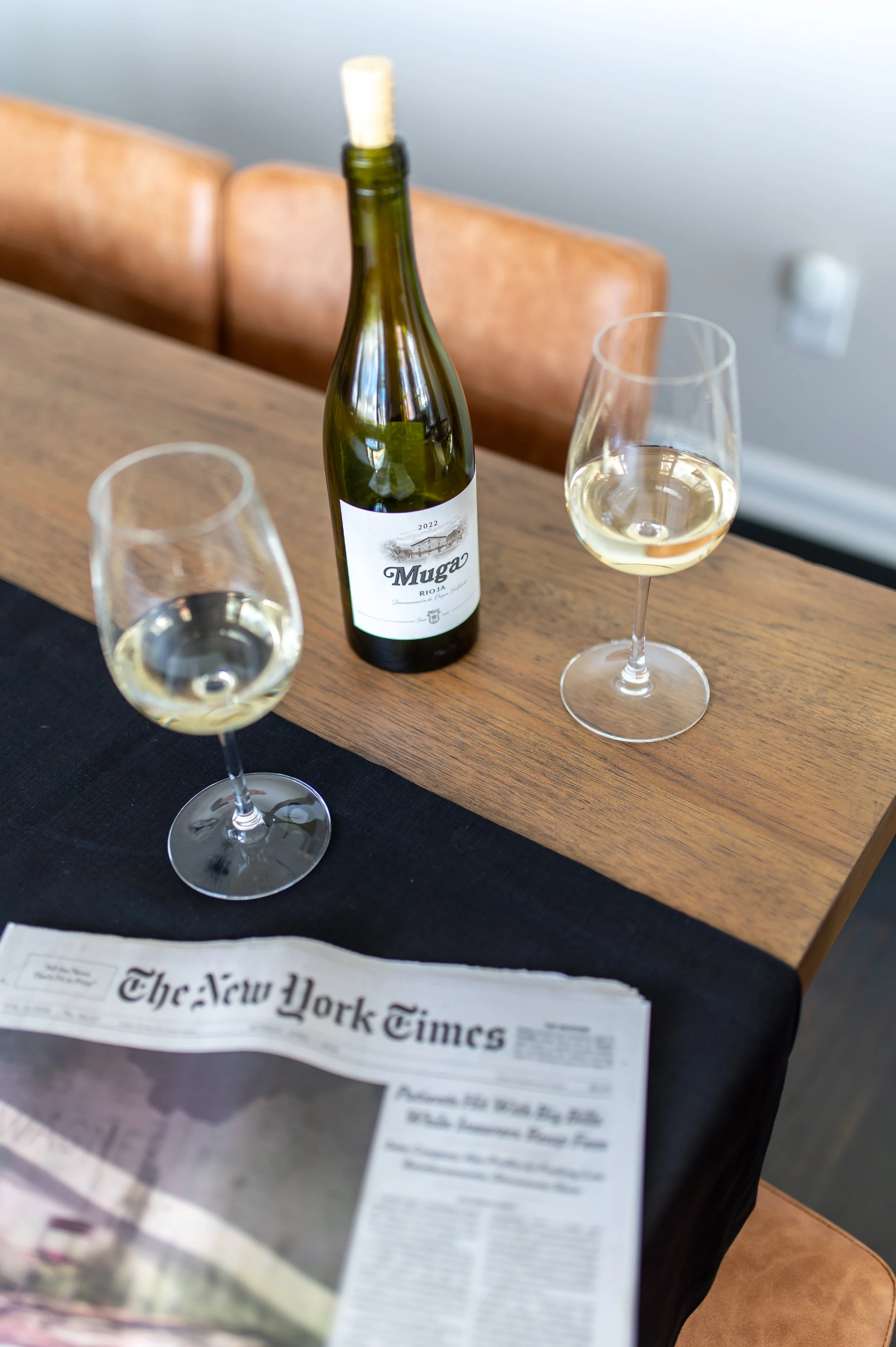A bottle of white wine, two glasses of white wine, and a newspaper on a table.