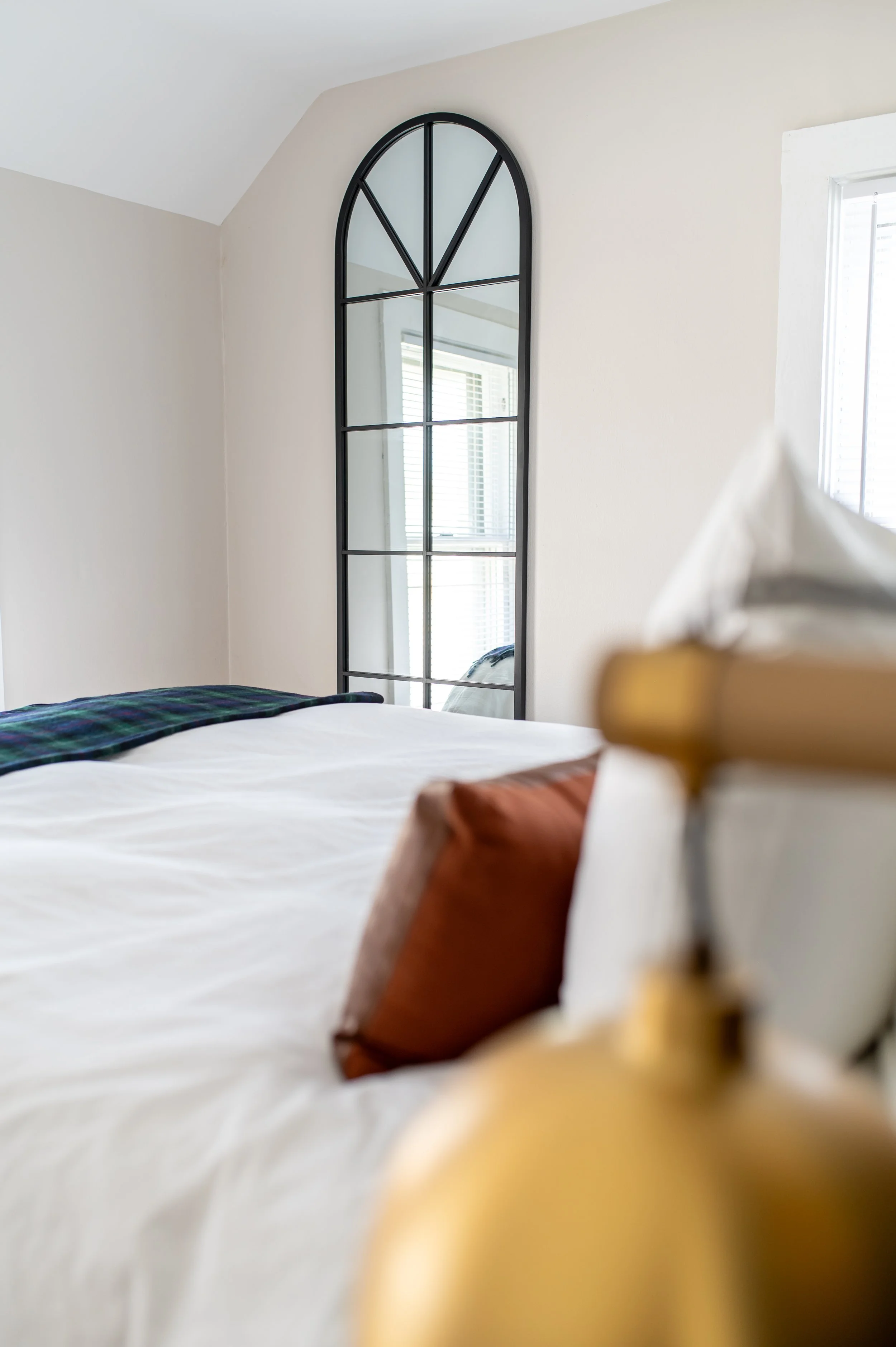 Close-up of a bed with white bedding and a brown pillow, with a gold-colored bedside lamp in the foreground, and a tall black-framed mirror and window with blinds in the background.