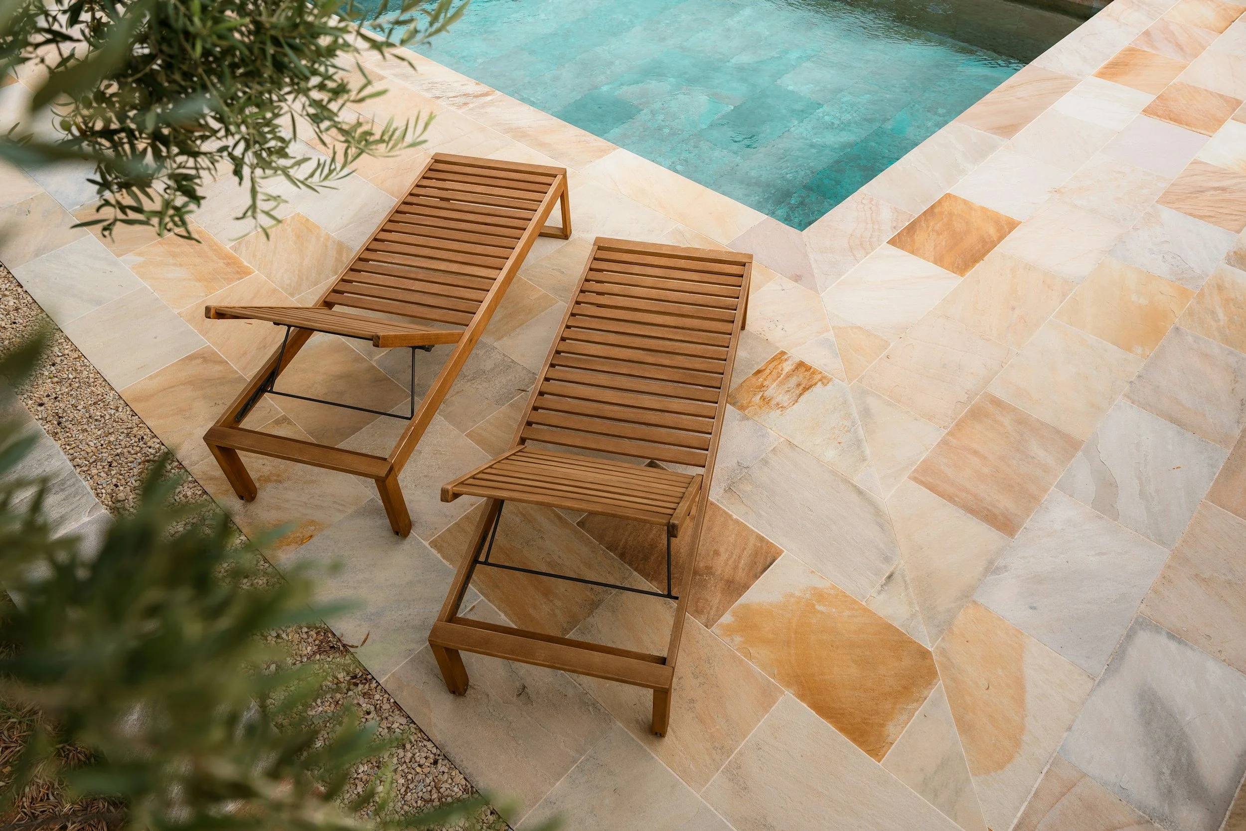 Two wooden lounge chairs on a stone patio next to a swimming pool.