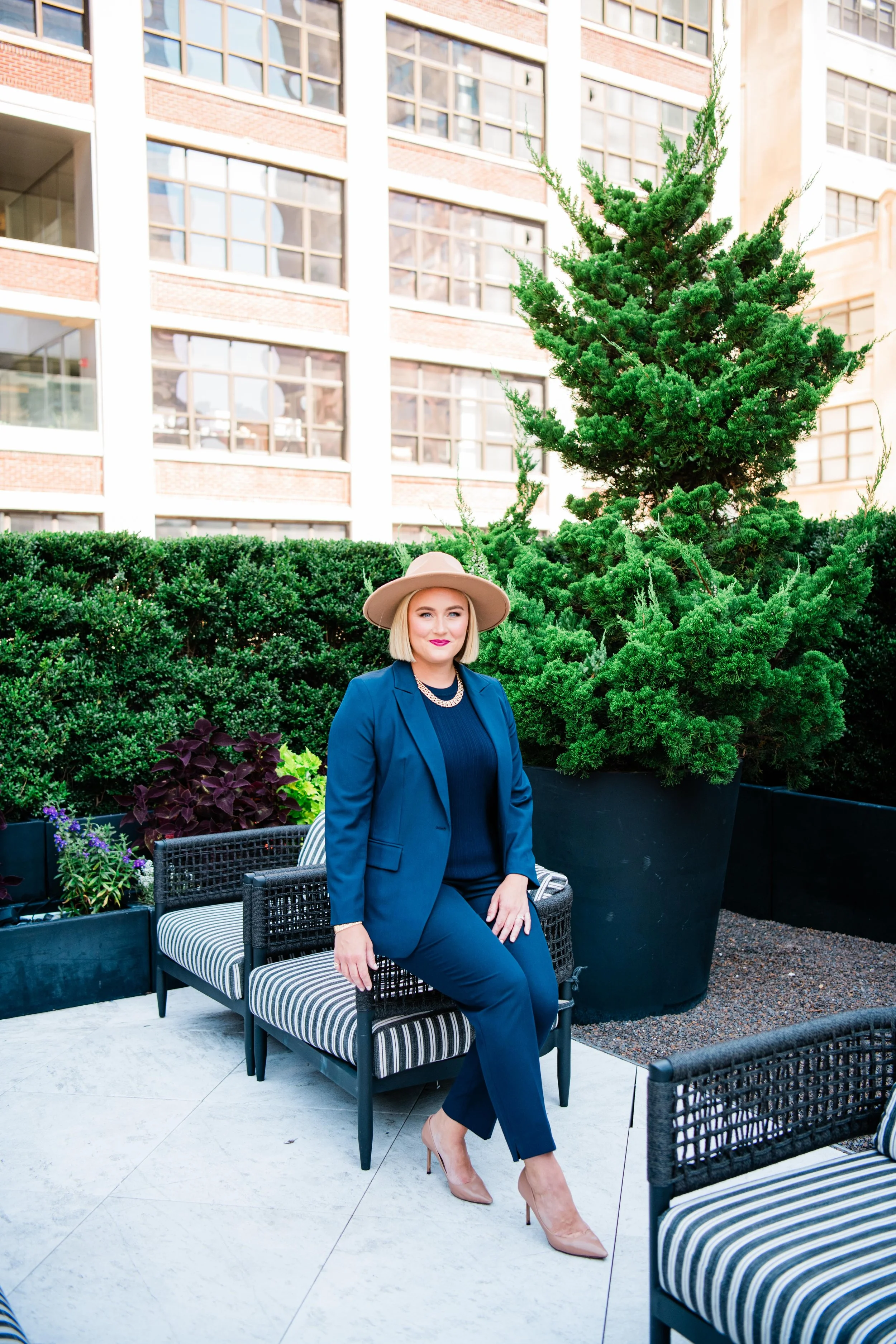 A woman in a navy blue suit and beige heels sitting outdoors on a striped bench with her hand resting on her knee, wearing a beige wide-brimmed hat and jewelry, with a large potted evergreen tree behind her and a multi-story building in the background.