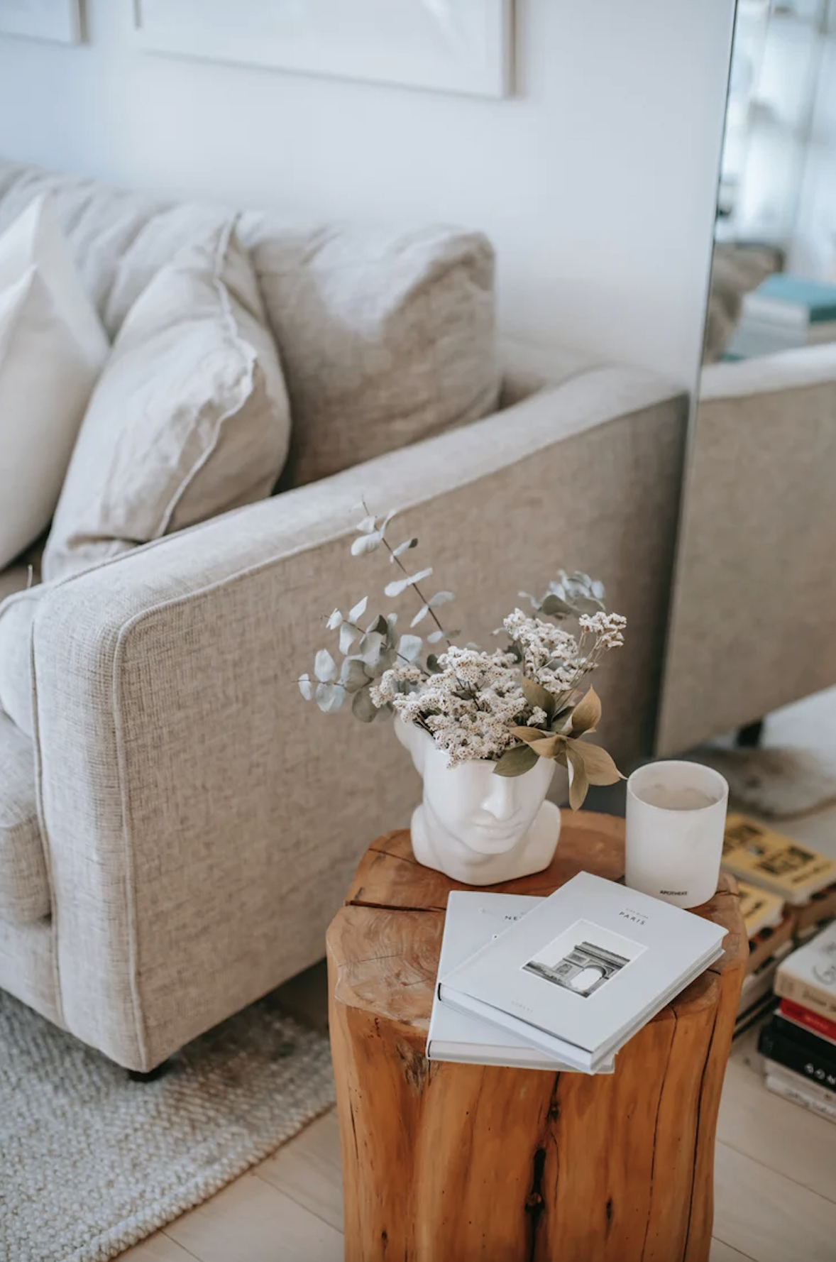 A wooden side table holding a white decorative head-shaped vase with dried flowers, a white candle, and two books, in front of a beige upholstered sofa with cushions in a cozy living room.