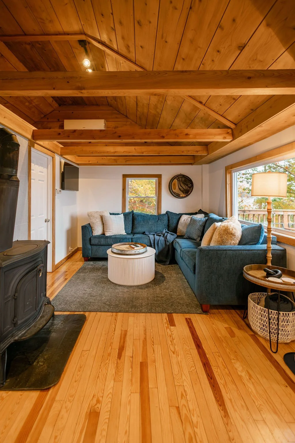 Living room with a wooden ceiling, blue sectional sofa with pillows, window showing autumn trees, wood floor, and a wood stove.