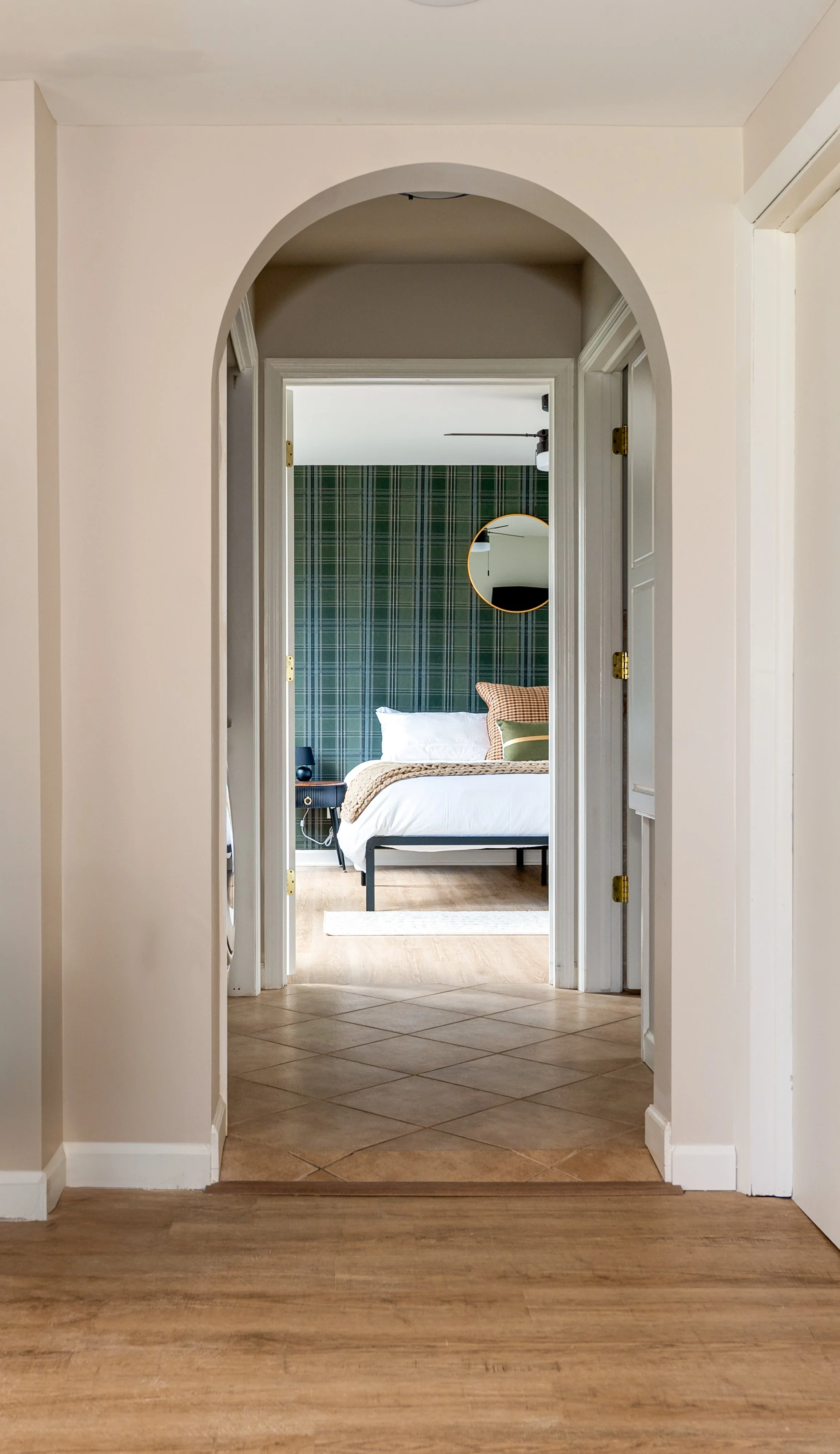 View through an arched doorway into a bedroom with a bed, pillows, a round mirror on a wall with green plaid wallpaper, and a wooden floor.