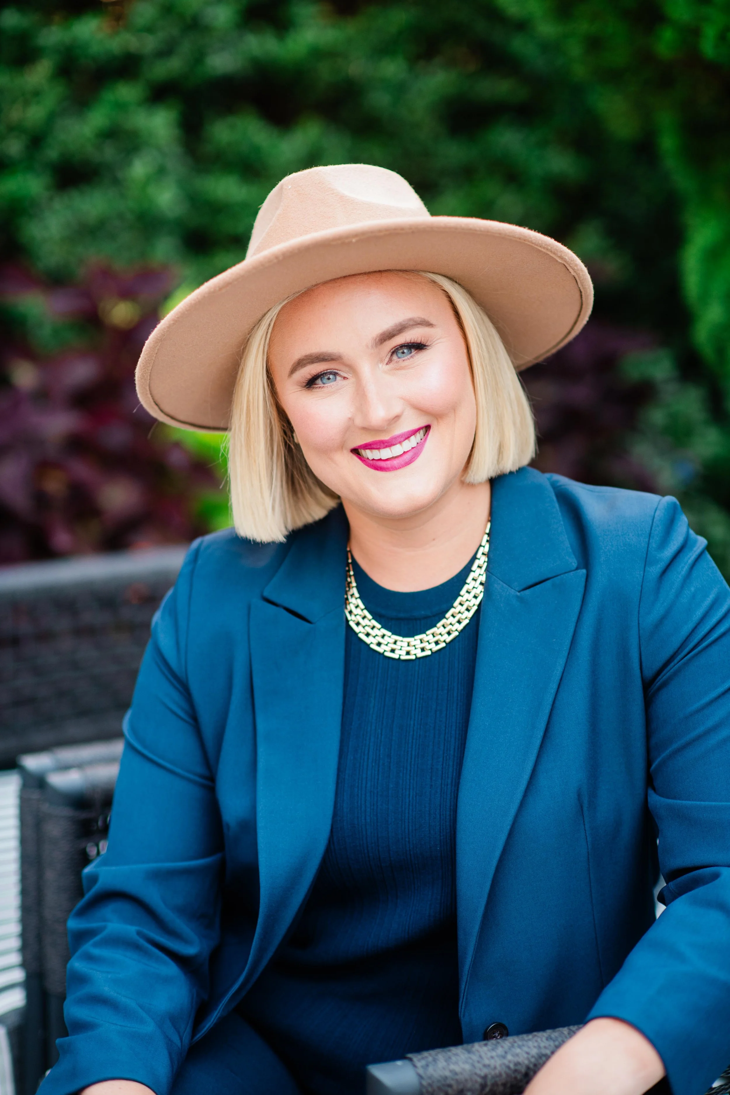 A smiling woman with blonde hair wearing a tan wide-brim hat, blue blazer, and gold necklace, sitting outdoors with green foliage in the background.