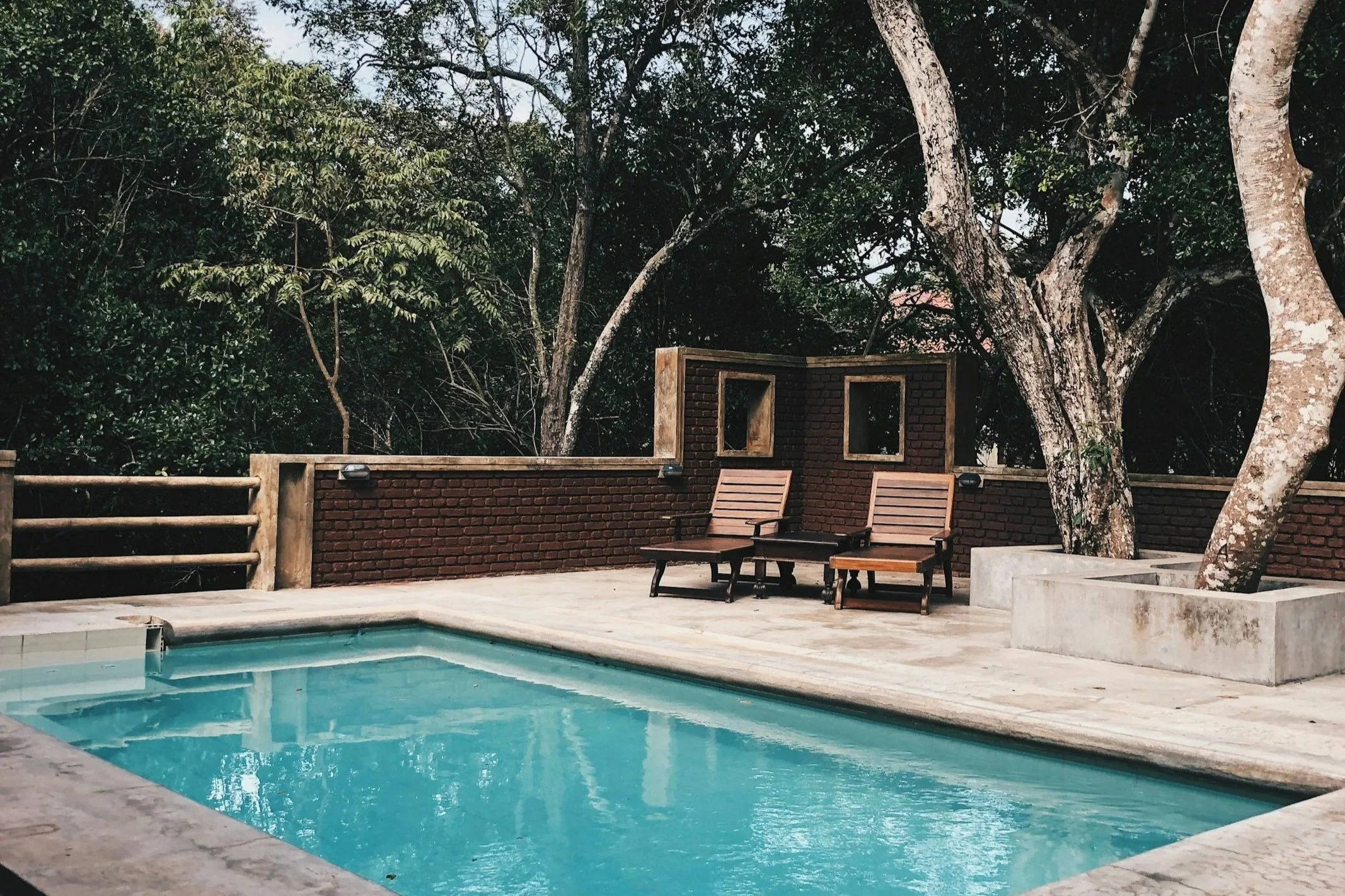 A backyard swimming pool with two lounge chairs and a small table, surrounded by trees and a brick fence.