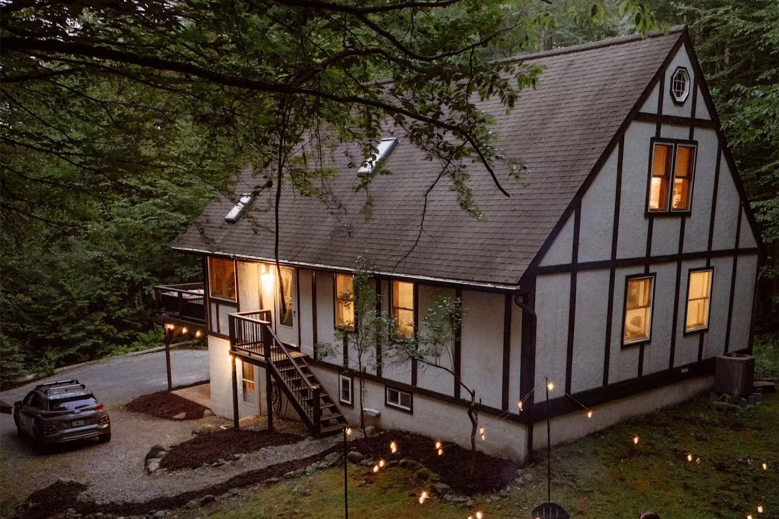 A two-story house with black and white exterior, large windows with warm light inside, a staircase leading to the front door, surrounded by trees, with string lights in the yard and a black vehicle parked nearby