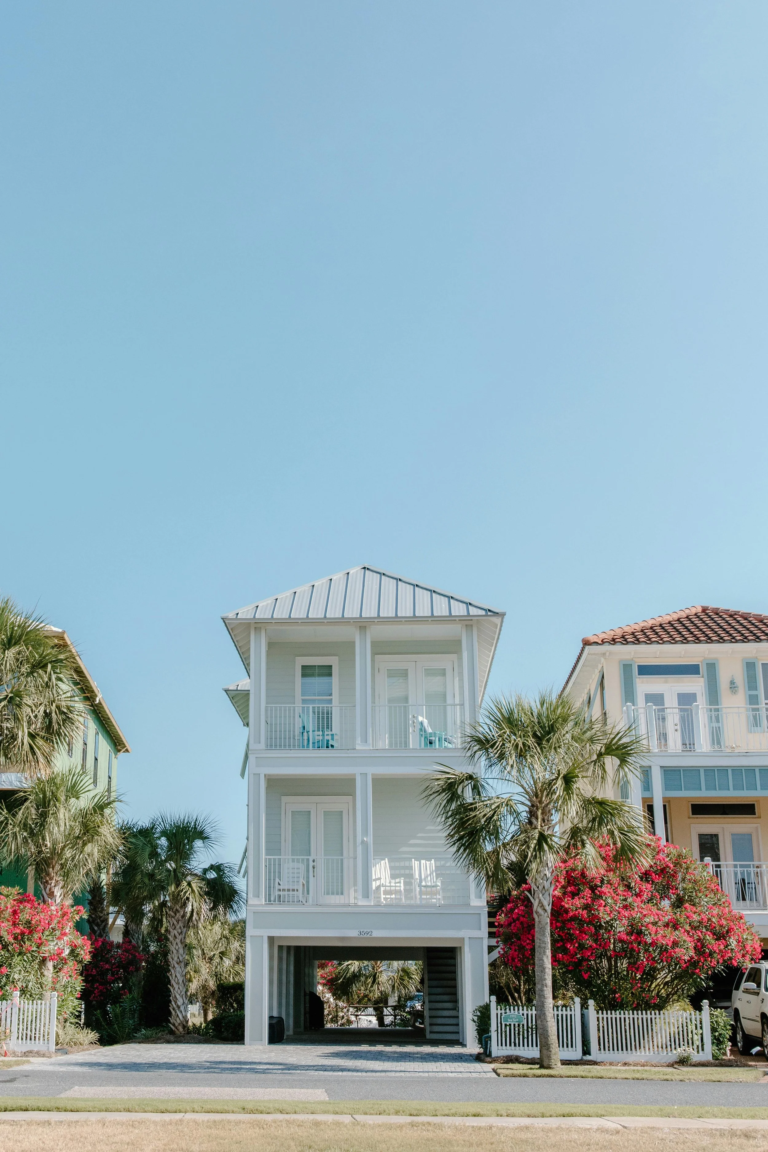 A three-story beach house painted white with balconies on each floor, set amidst palm trees and pink flowering bushes, under a clear blue sky.