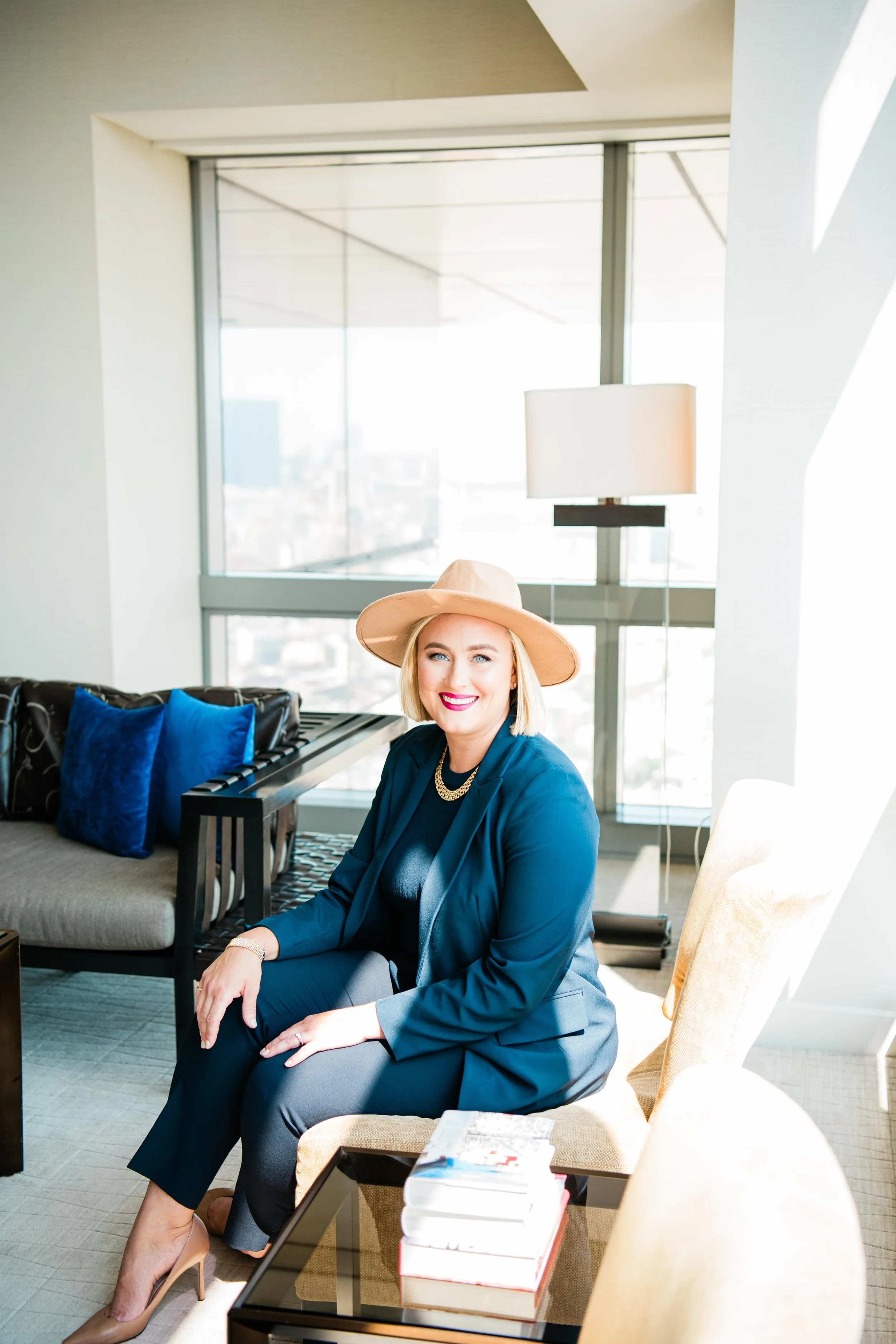 A woman with blonde hair wearing a beige hat, blue blazer, black top, and grey pants, sitting on a beige chair in a bright, modern room with large windows.