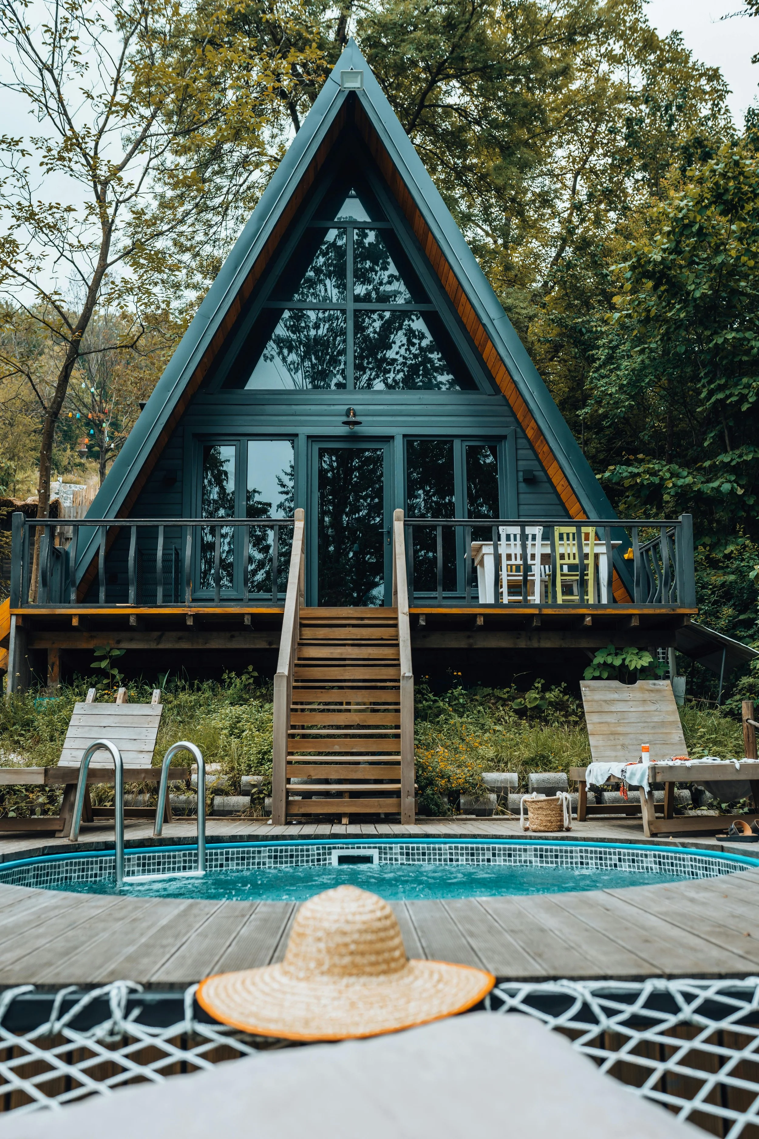 A-frame cabin with large glass windows and wooden deck, surrounded by trees, overlooking a swimming pool with lounge chairs and a straw hat in the foreground.