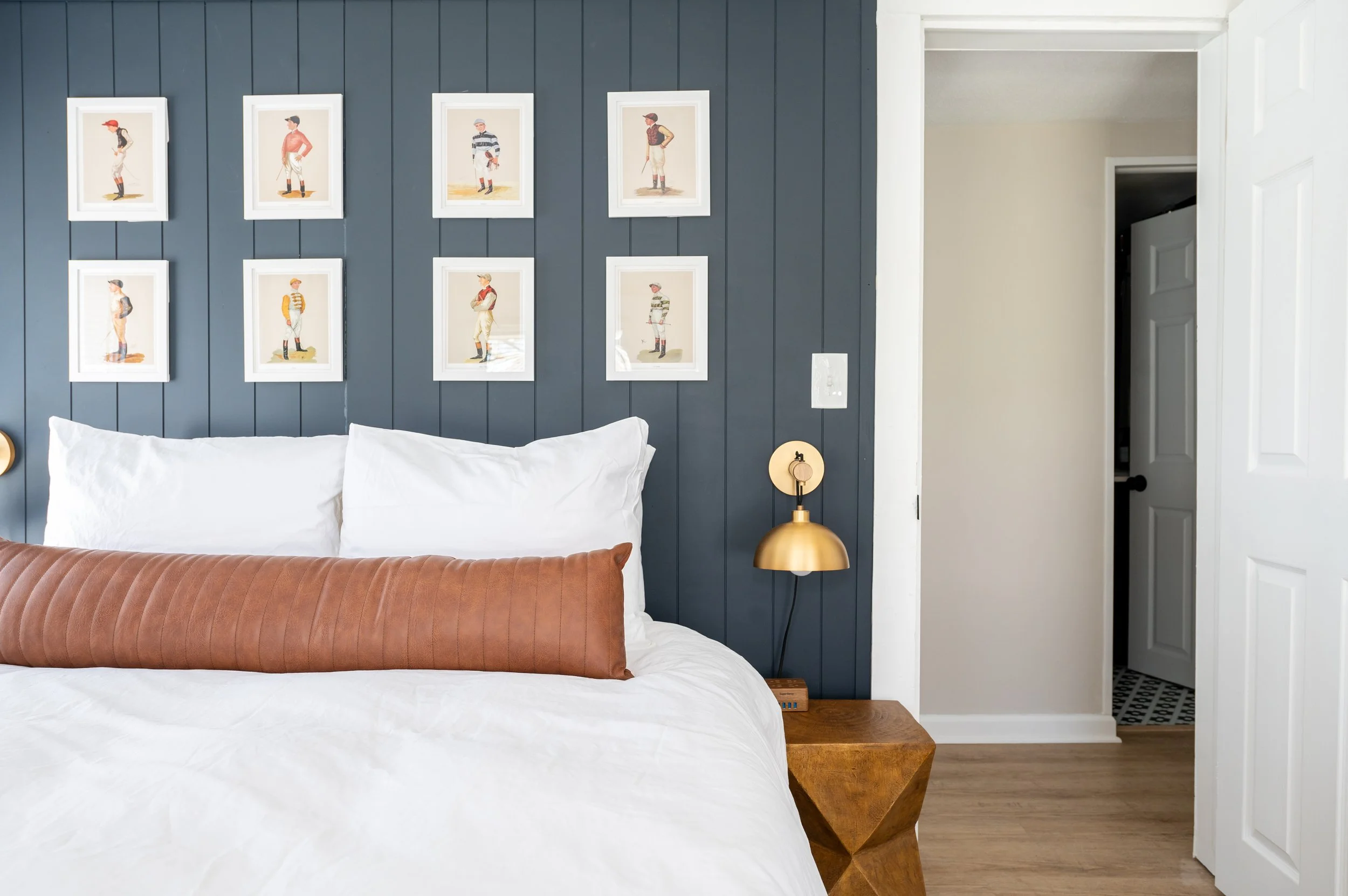 Modern bedroom with a navy blue accent wall decorated with framed illustrations of children in nautical outfits. A bed with white bedding and a long brown pillow is positioned in front of the wall, with a wooden nightstand and a gold wall-mounted lamp to the right of the bed. An open door reveals a glimpse of a hallway.