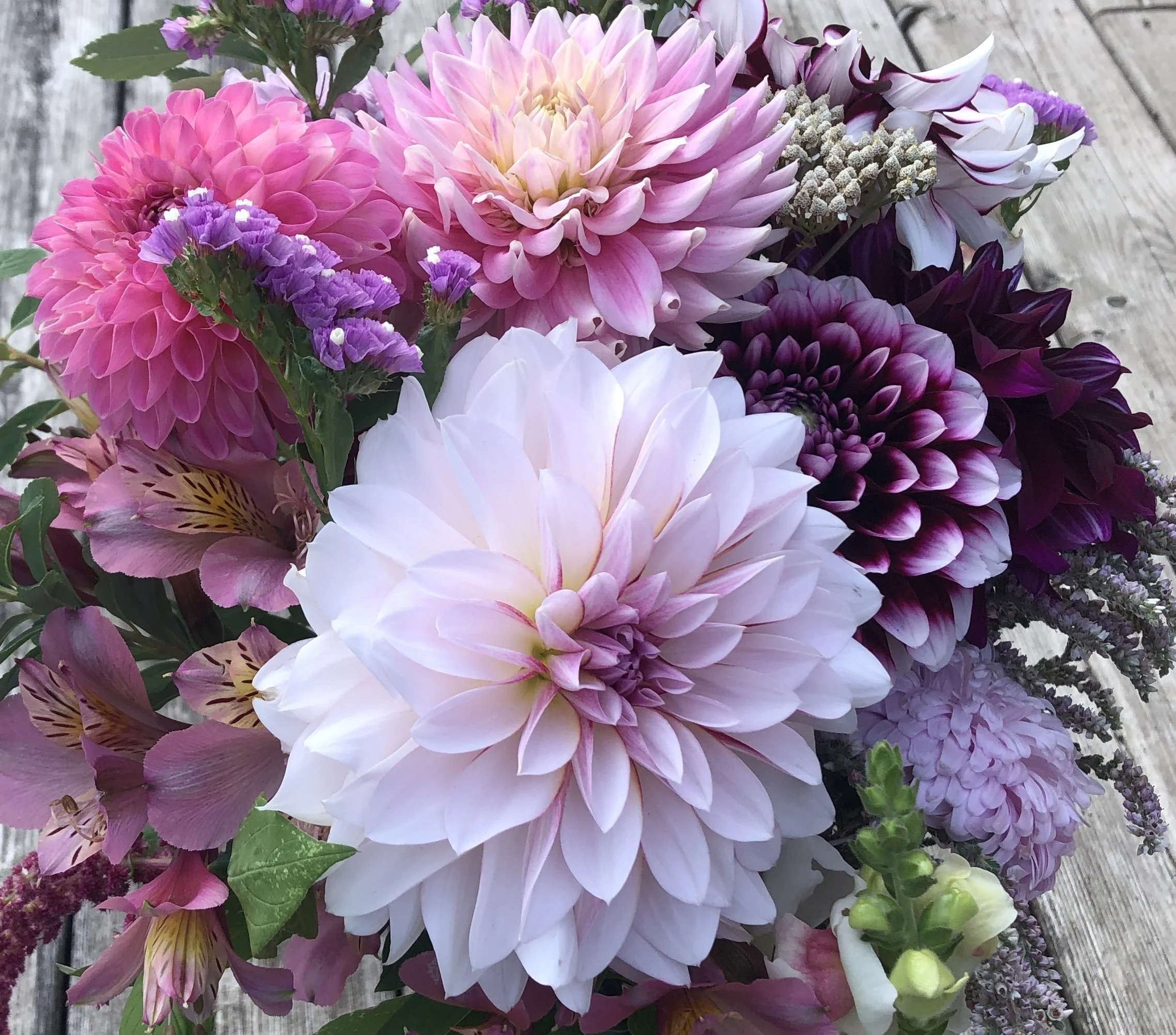 A bouquet of pink, purple, and white flowers, including dahlias, roses, and other mixed blooms, arranged on a wooden surface.