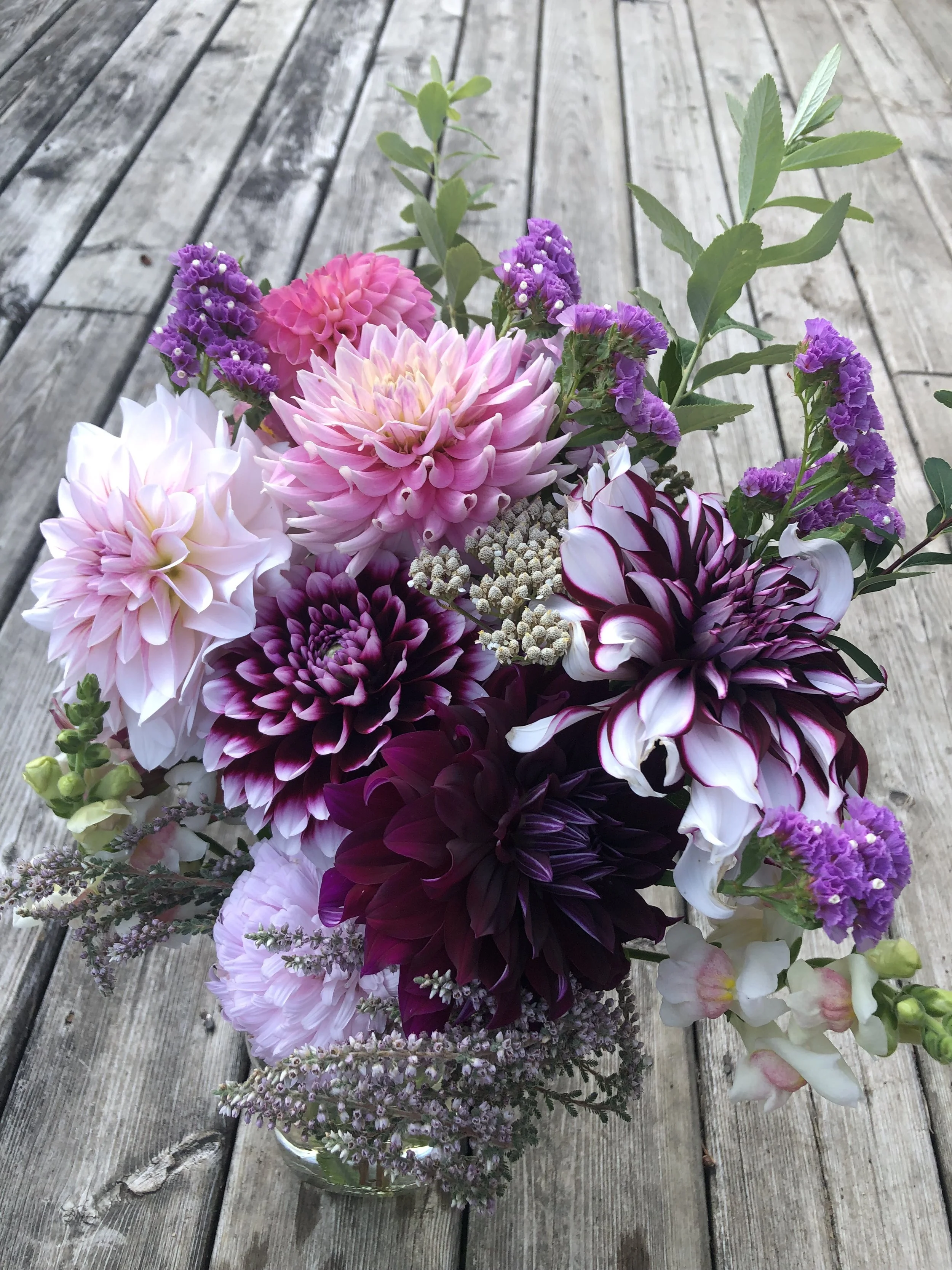A vibrant bouquet of various colorful dahlias and other flowers in a glass jar on a wooden surface.