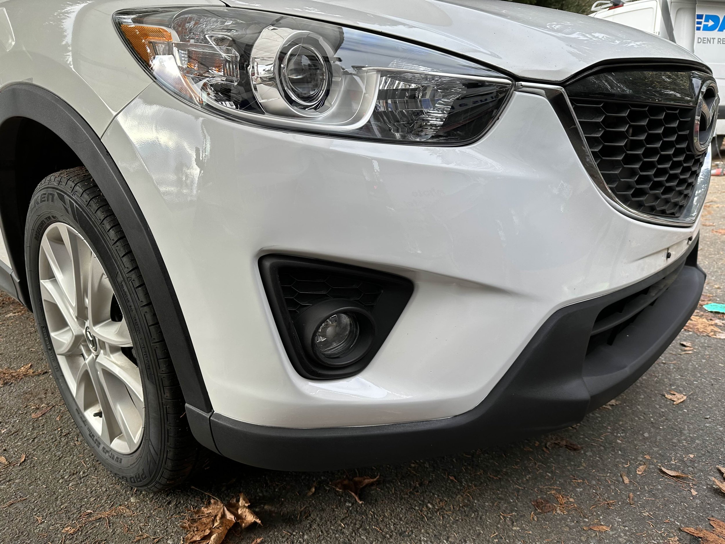Close-up of the front left side of a white Mazda SUV, showing the headlight, tire, black front grille, and fog light with a black bumper.