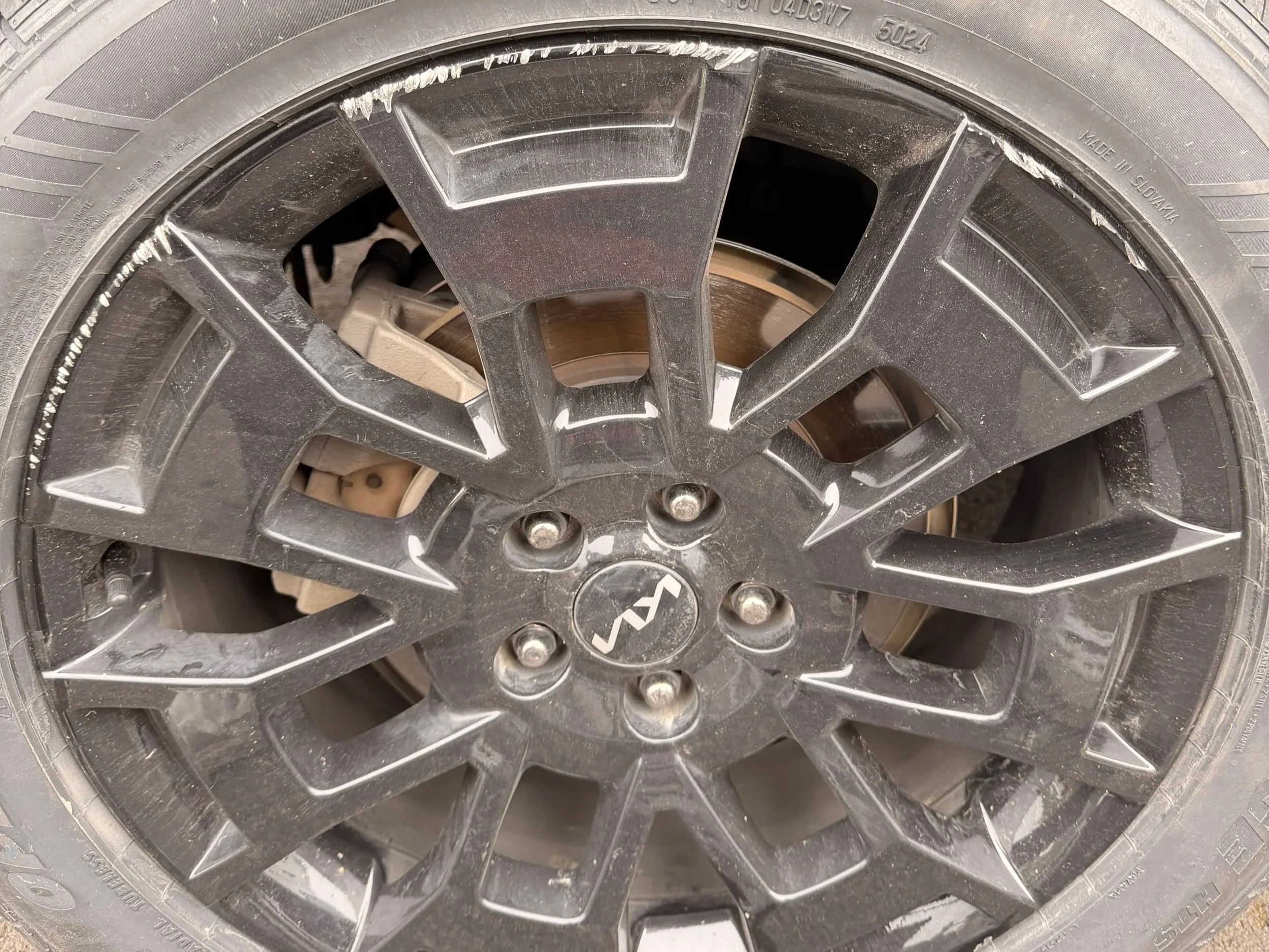 Close-up of a black alloy wheel with a logo at the center, dusty and with some white marks on the rim, covered by a tire.