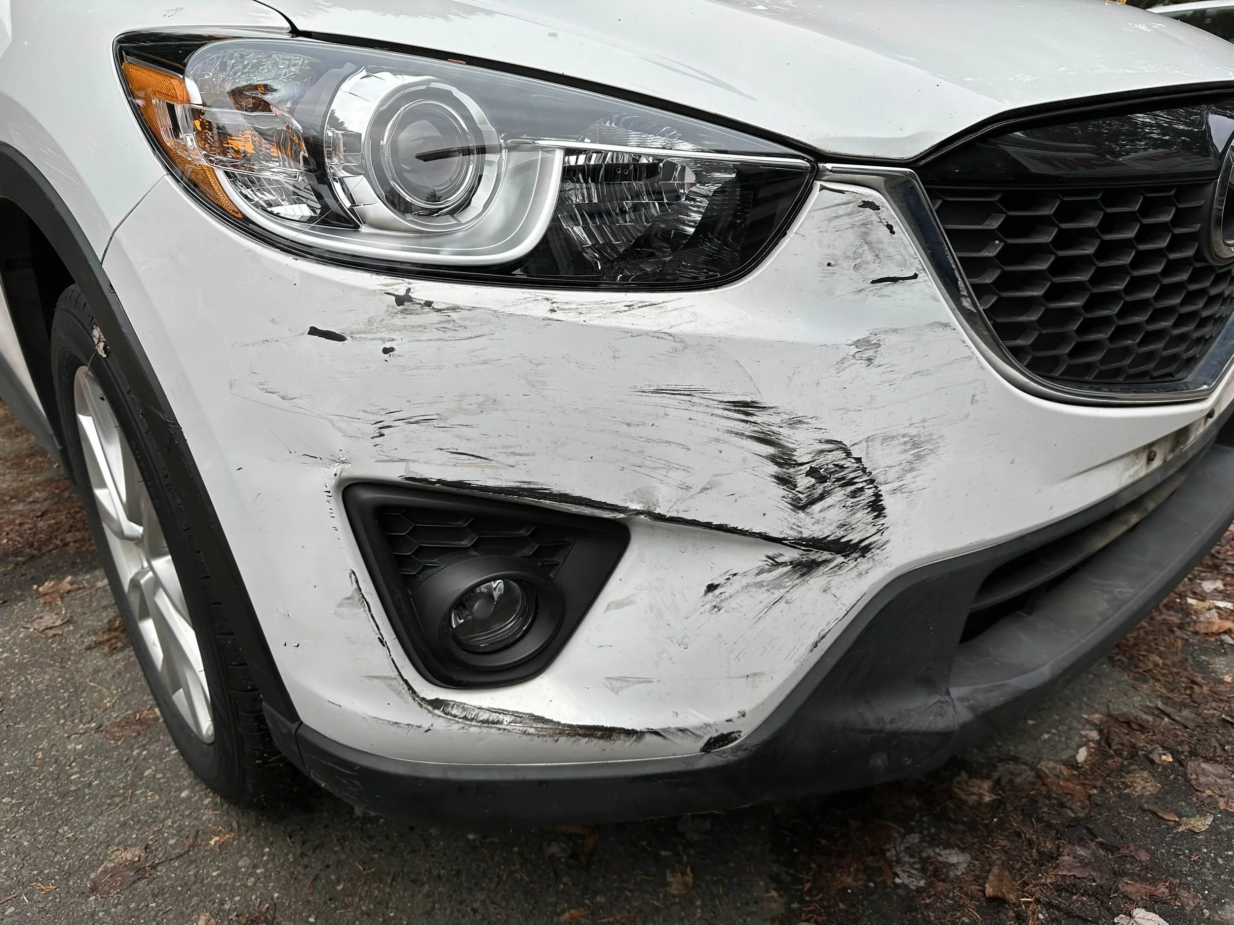Front of a silver SUV with visible front-end damage, including scratches and dents on the left side of the bumper and fender, on a street with fallen leaves.