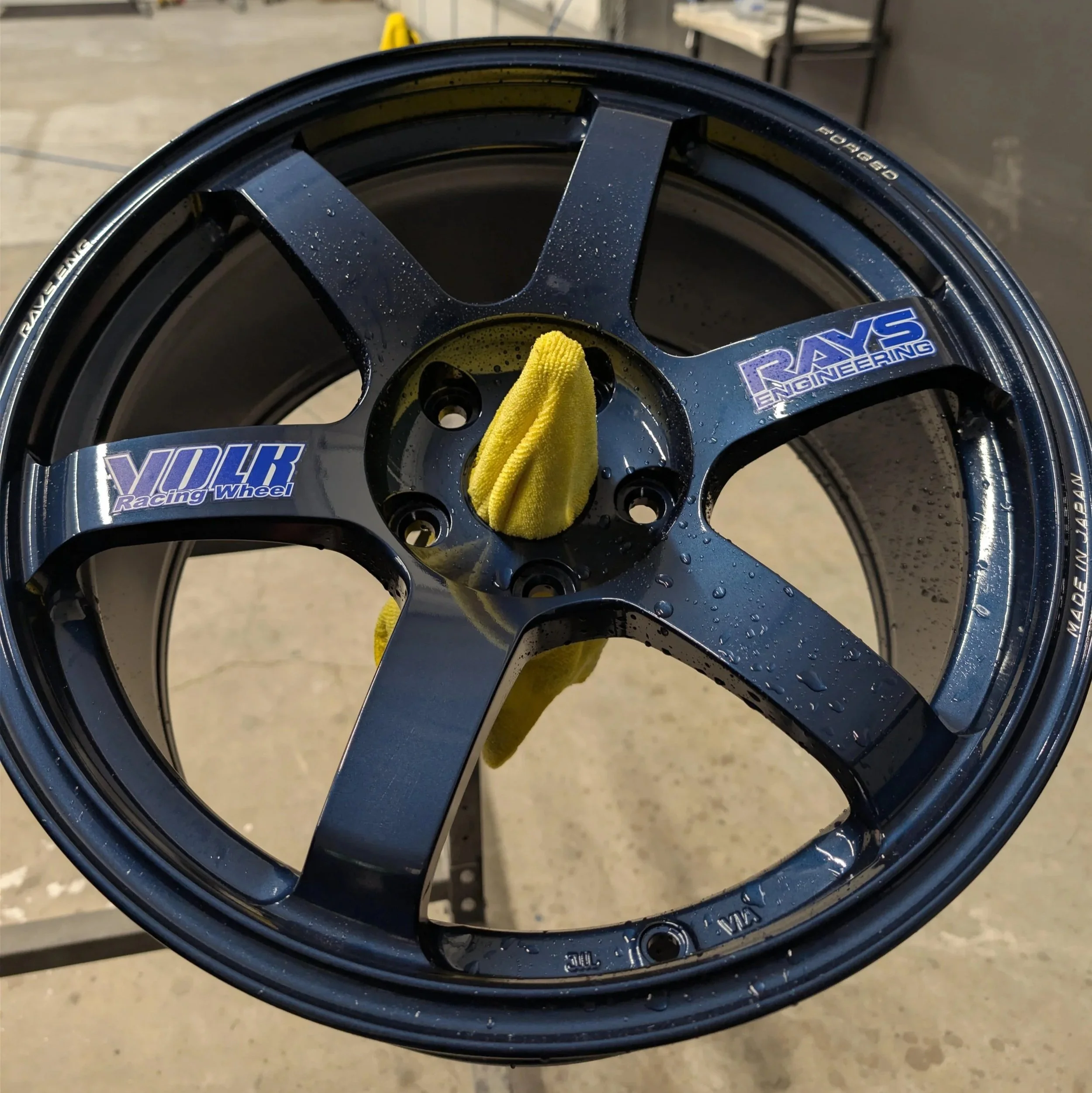 Close-up view of a black racing wheel rim with water droplets and a yellow cloth hanging through the center, labeled with "YOLK Racing Wheel" and a part number engraved along the edge.