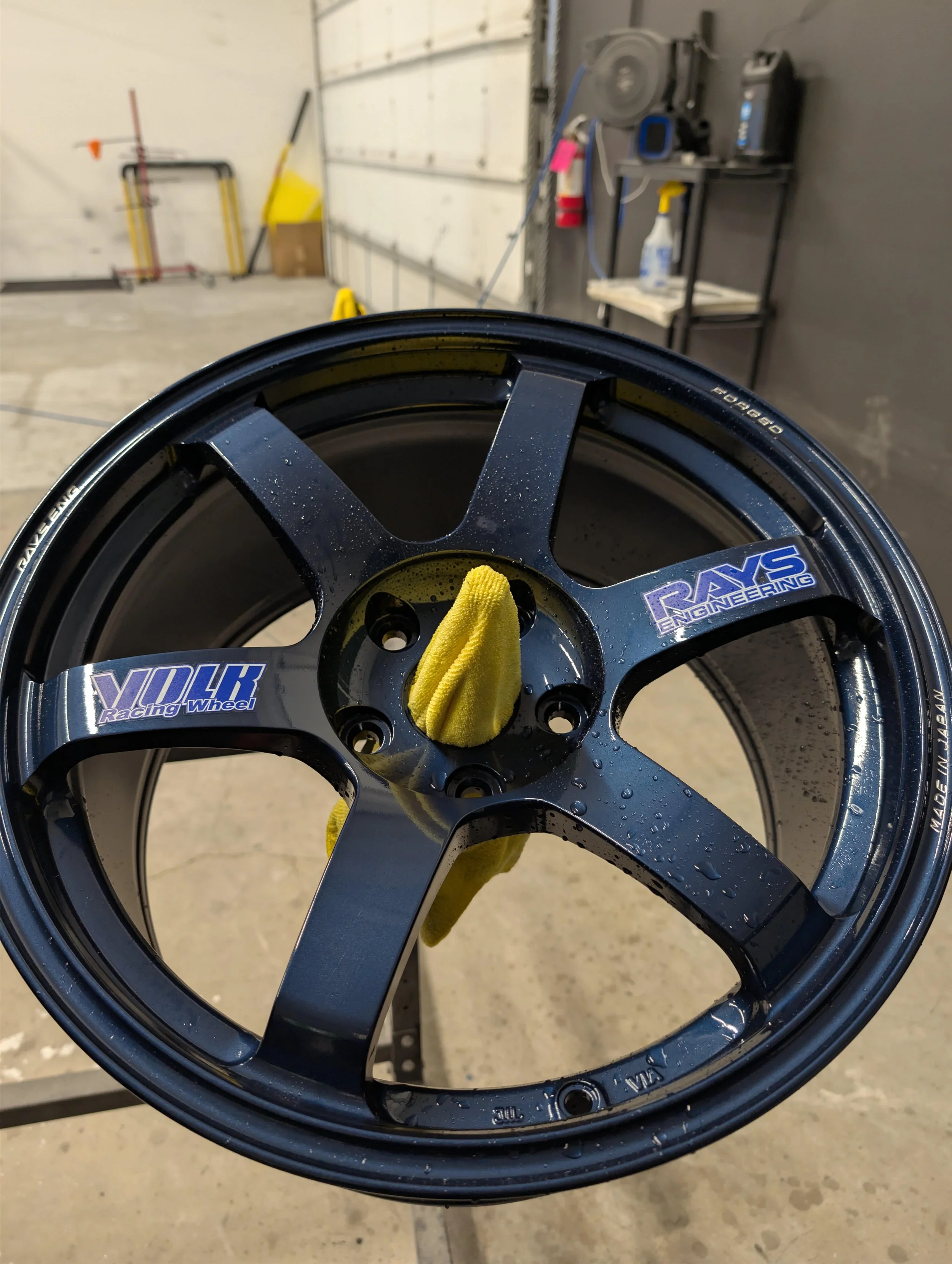 Close-up of a black racing wheel with a yellow cloth inside, water droplets on the surface, and a sticker that says 'VOLK RACING WHEEL.' The background shows a garage with various tools, a metal table, and a white wall.