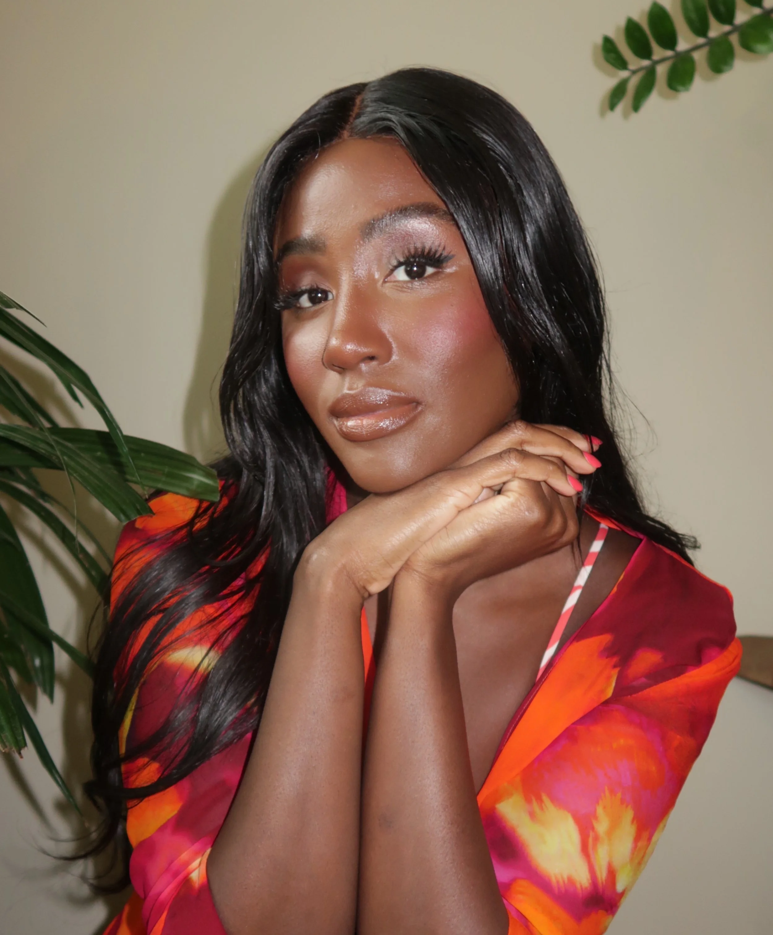 A woman with long black hair, wearing a colorful dress, is resting her chin on her hands near her face, surrounded by green leaves against a plain background.